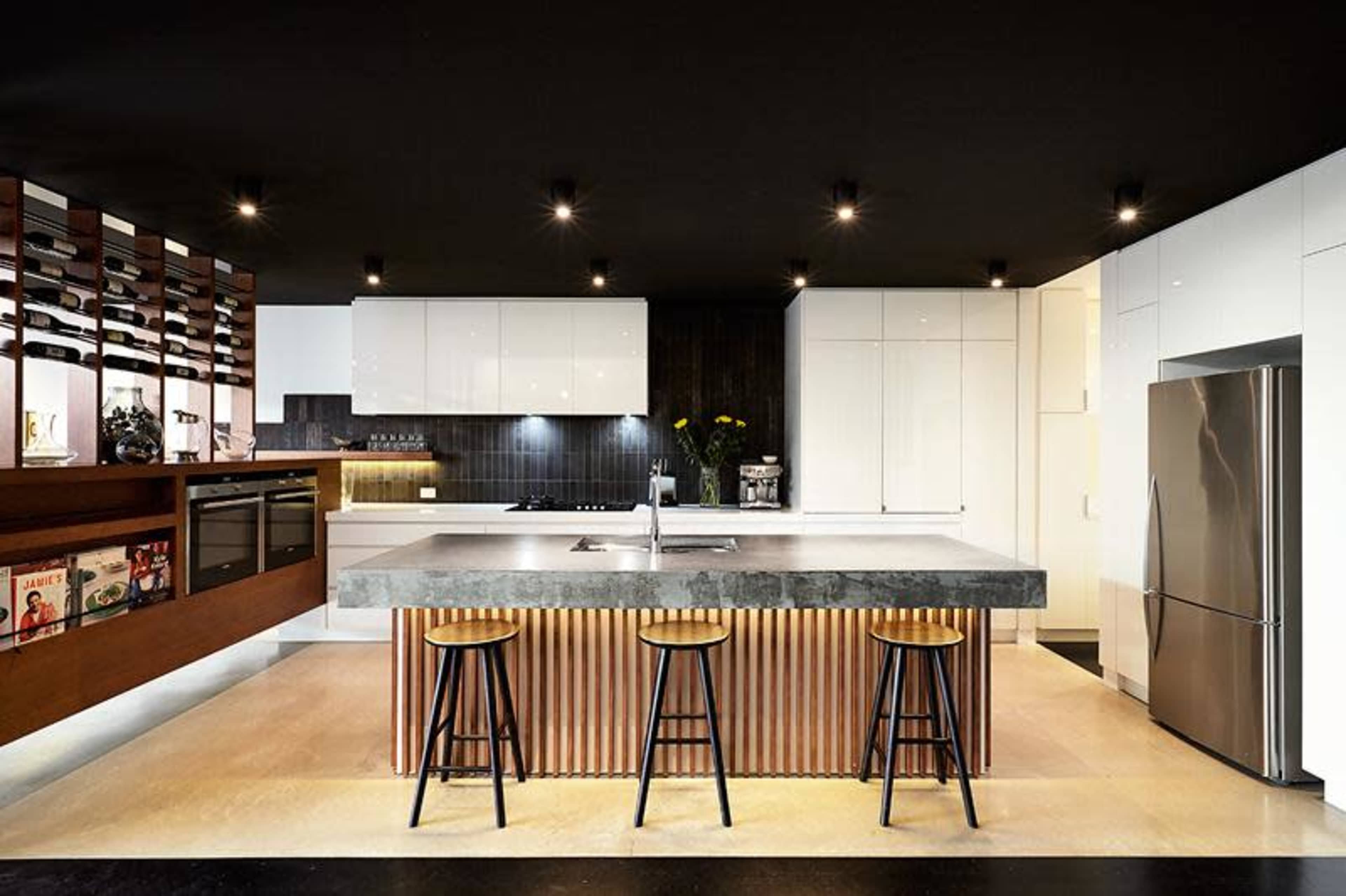 A modern kitchen features a central island with a stone countertop and metal stools, surrounded by sleek cabinetry and pendant lighting.