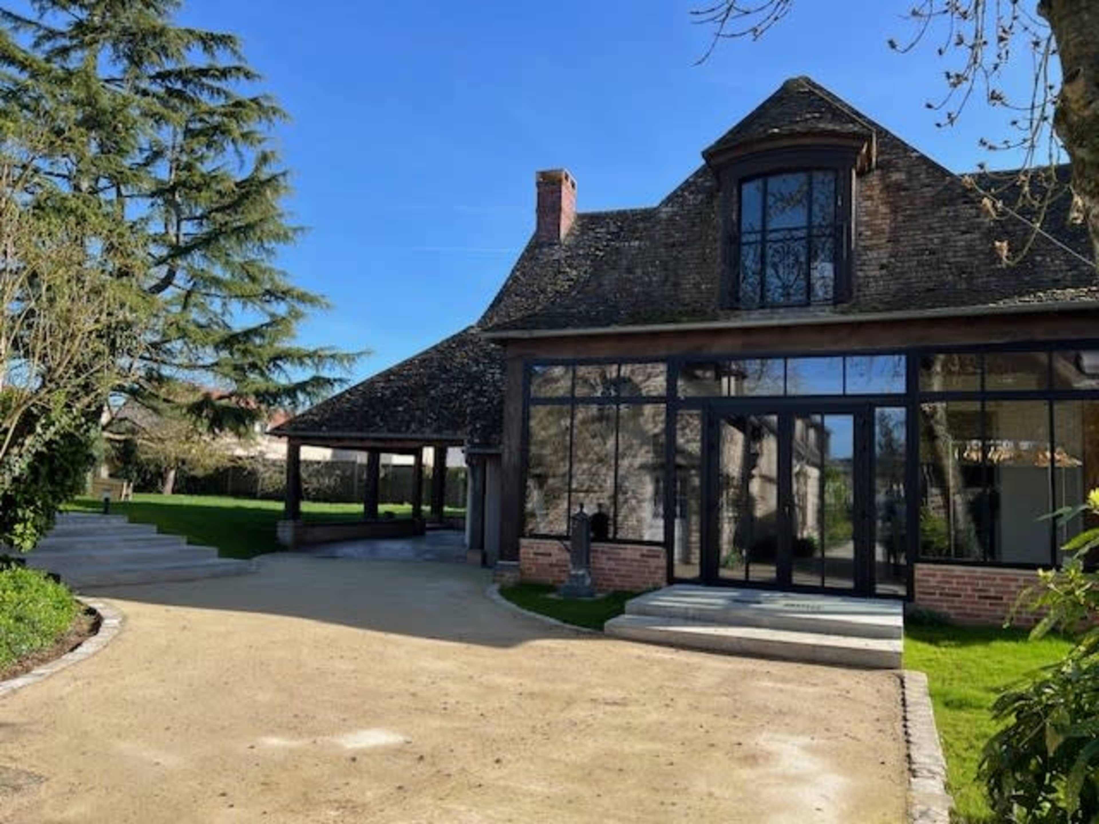 The image shows a modern house with large glass windows and a distinctive roof, situated in a landscaped area with trees and a gravel pathway leading up to it.