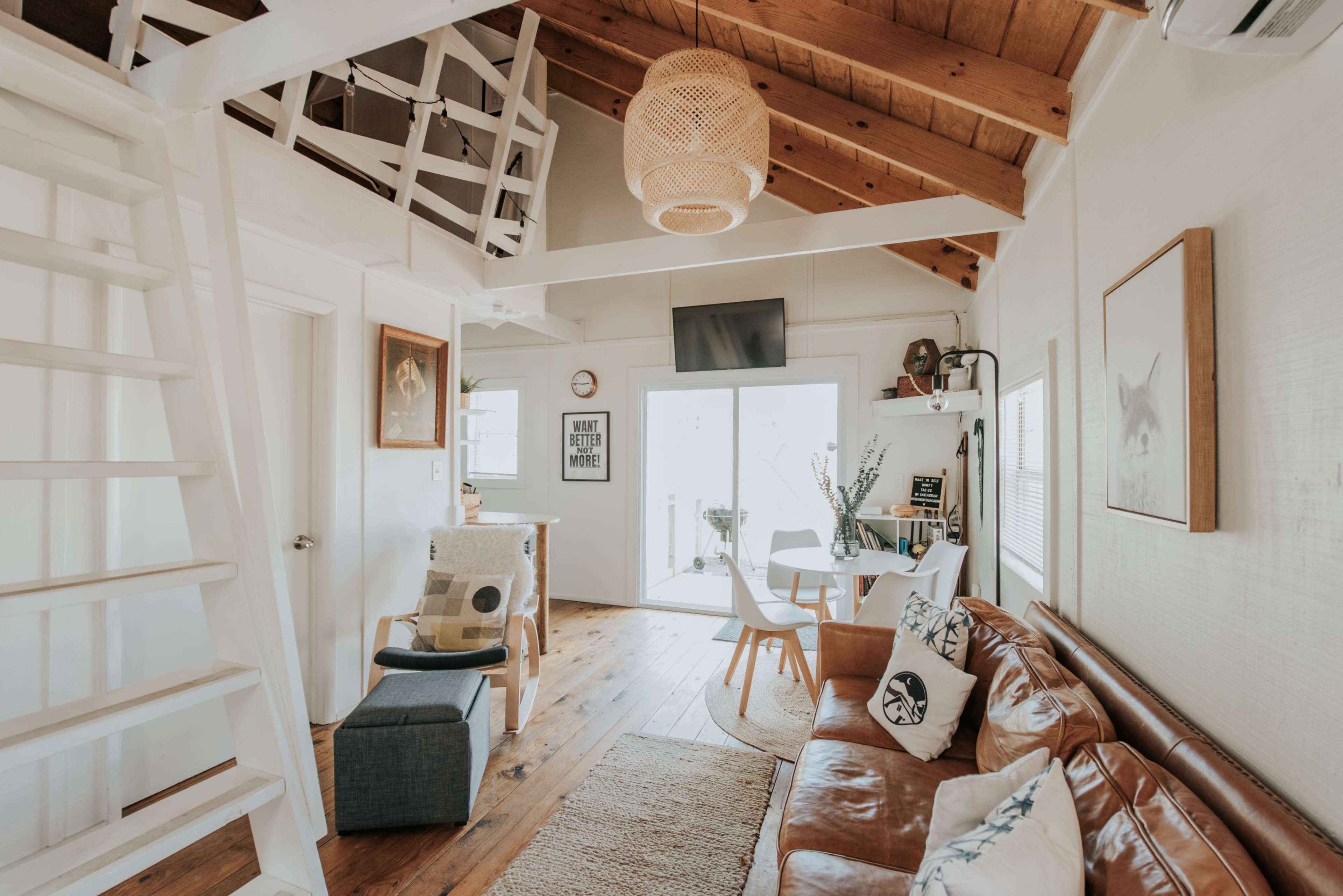 A cozy interior of a small living space featuring a brown leather sofa, a staircase leading to a loft, and a dining area with a round table and chairs.