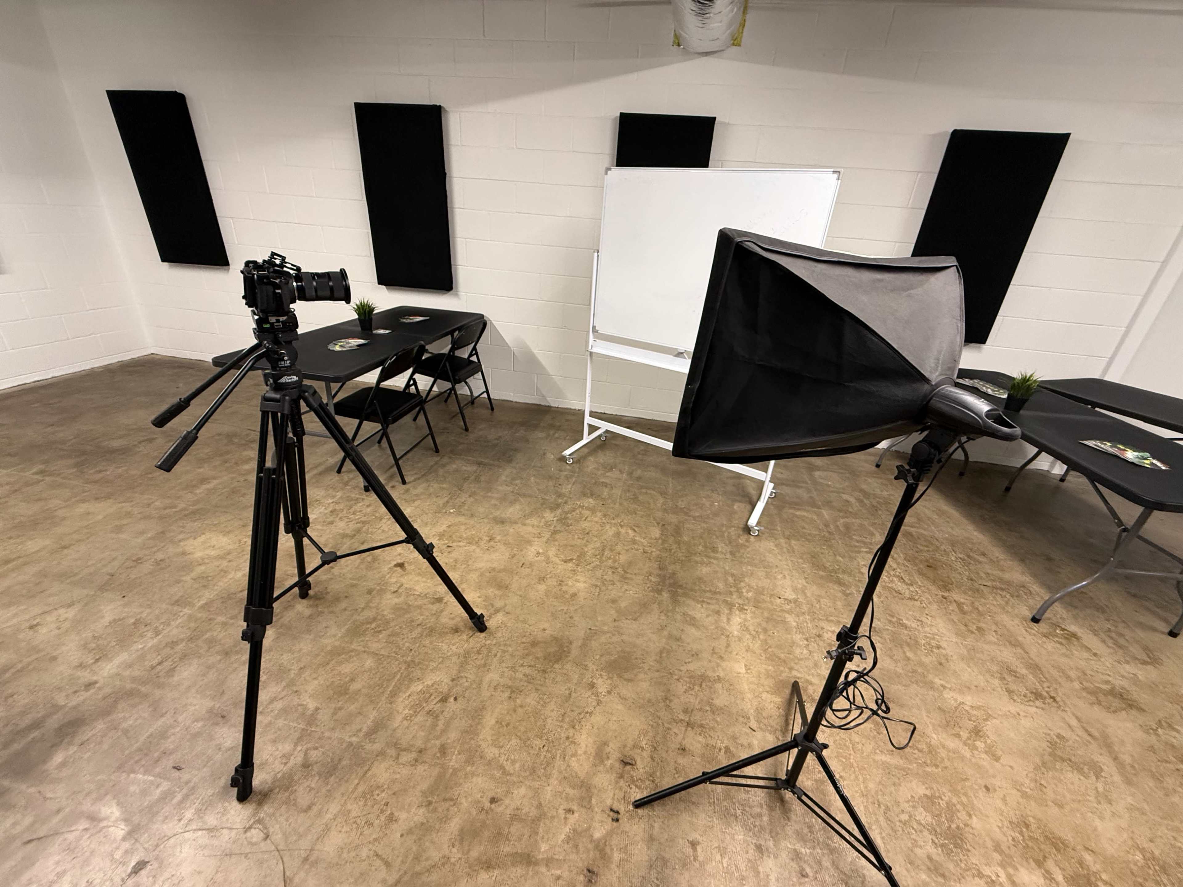 The image shows a photography setup in a room featuring a camera on a tripod, a softbox light, and a whiteboard against a wall.