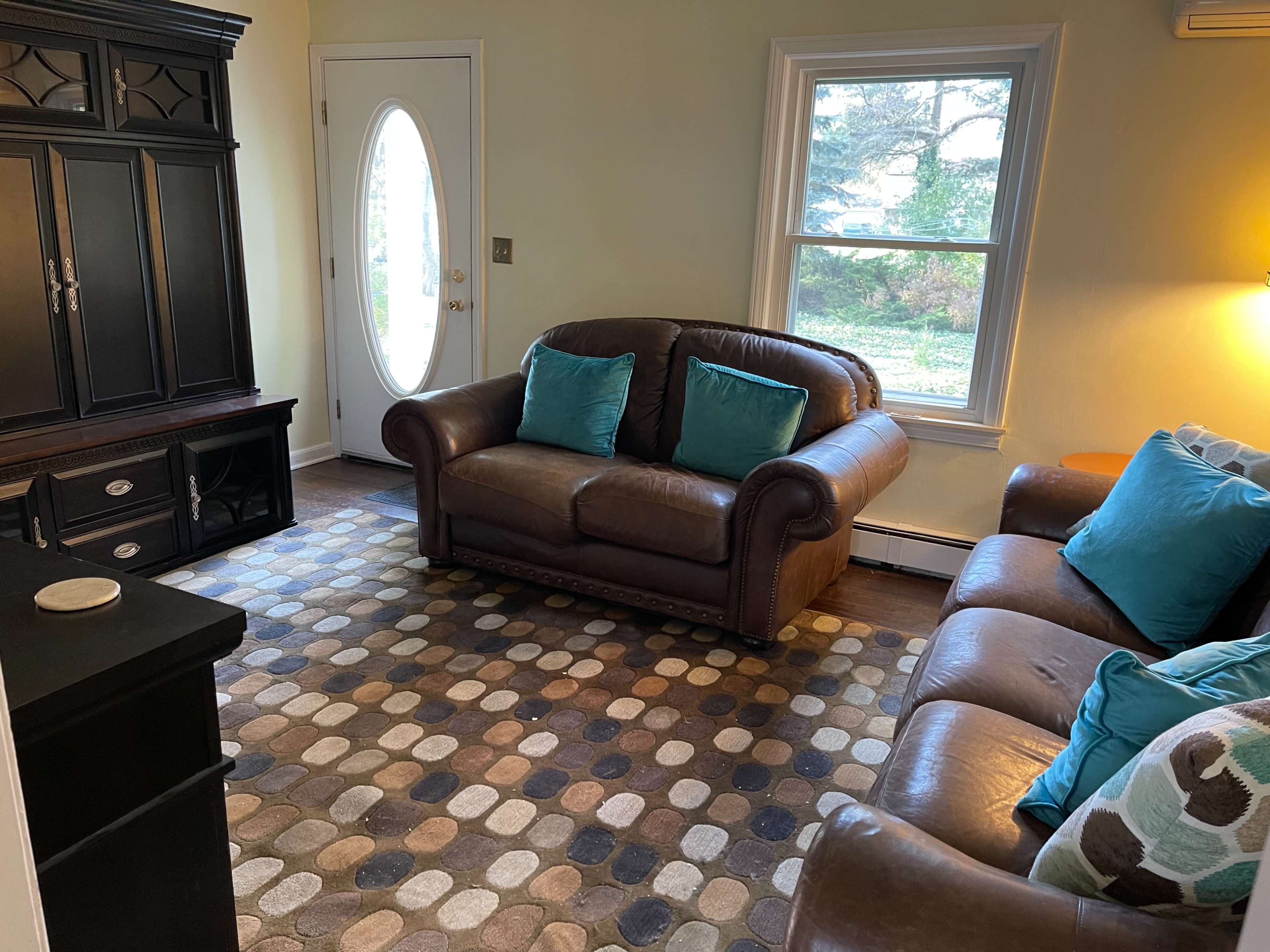 A living room features a brown leather sofa and loveseat, a circular patterned rug, and a door with a large round window allowing natural light.