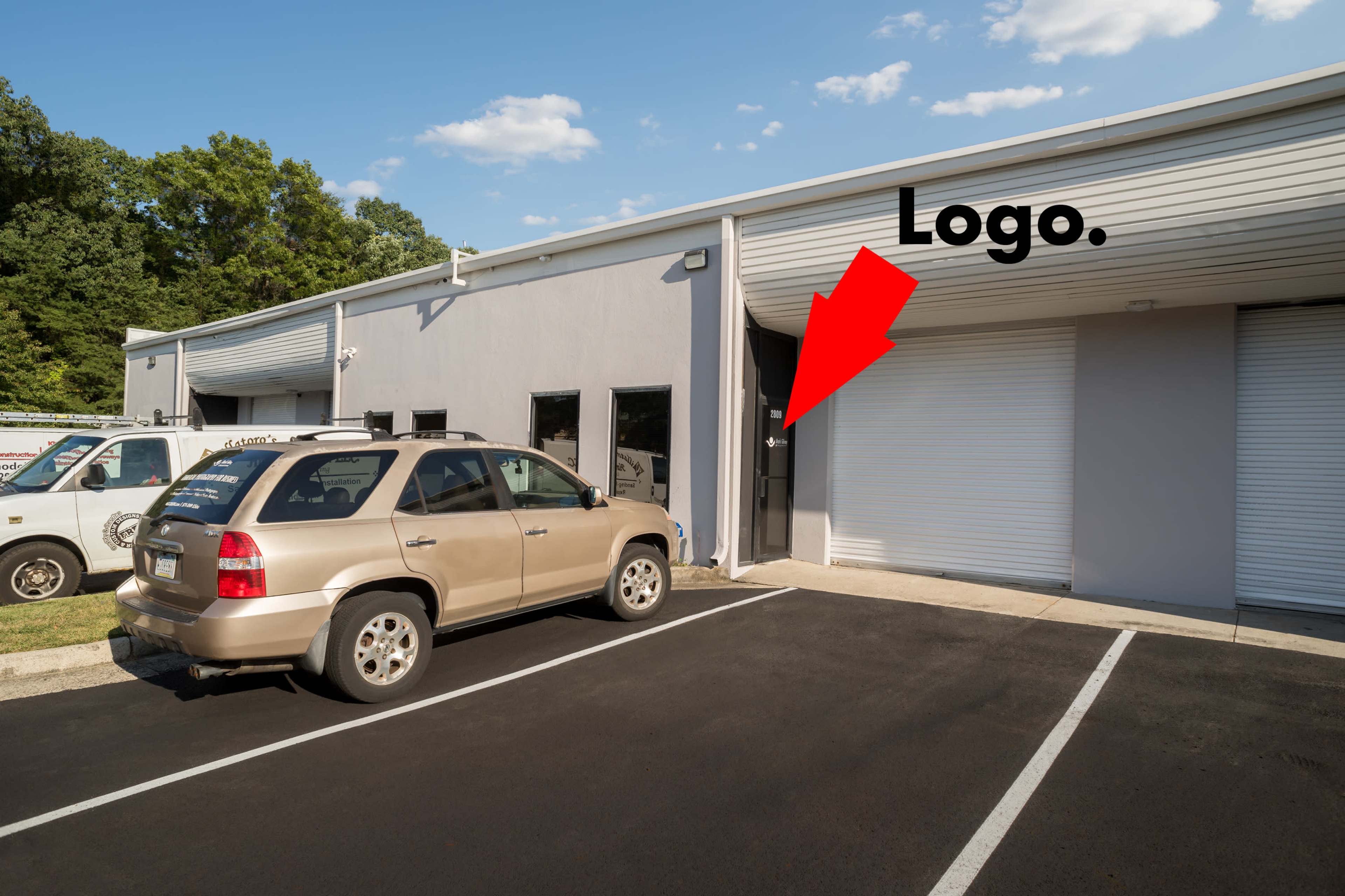 A beige SUV is parked in front of a commercial building with a gray facade, featuring a door marked with a logo and two large roll-up doors on the side.