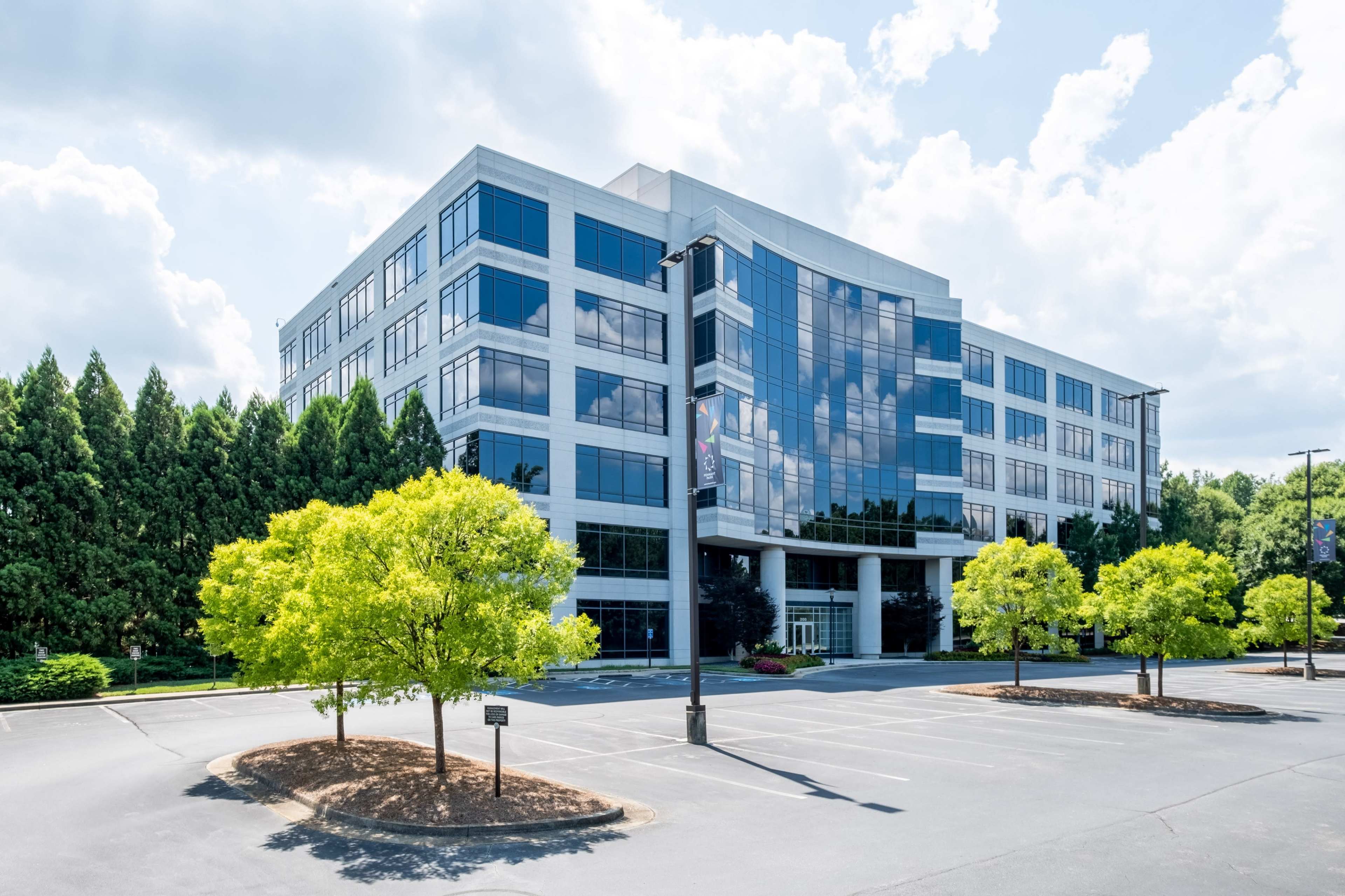 A modern office building with large windows is surrounded by neatly trimmed trees and an empty parking lot under a cloudy sky.