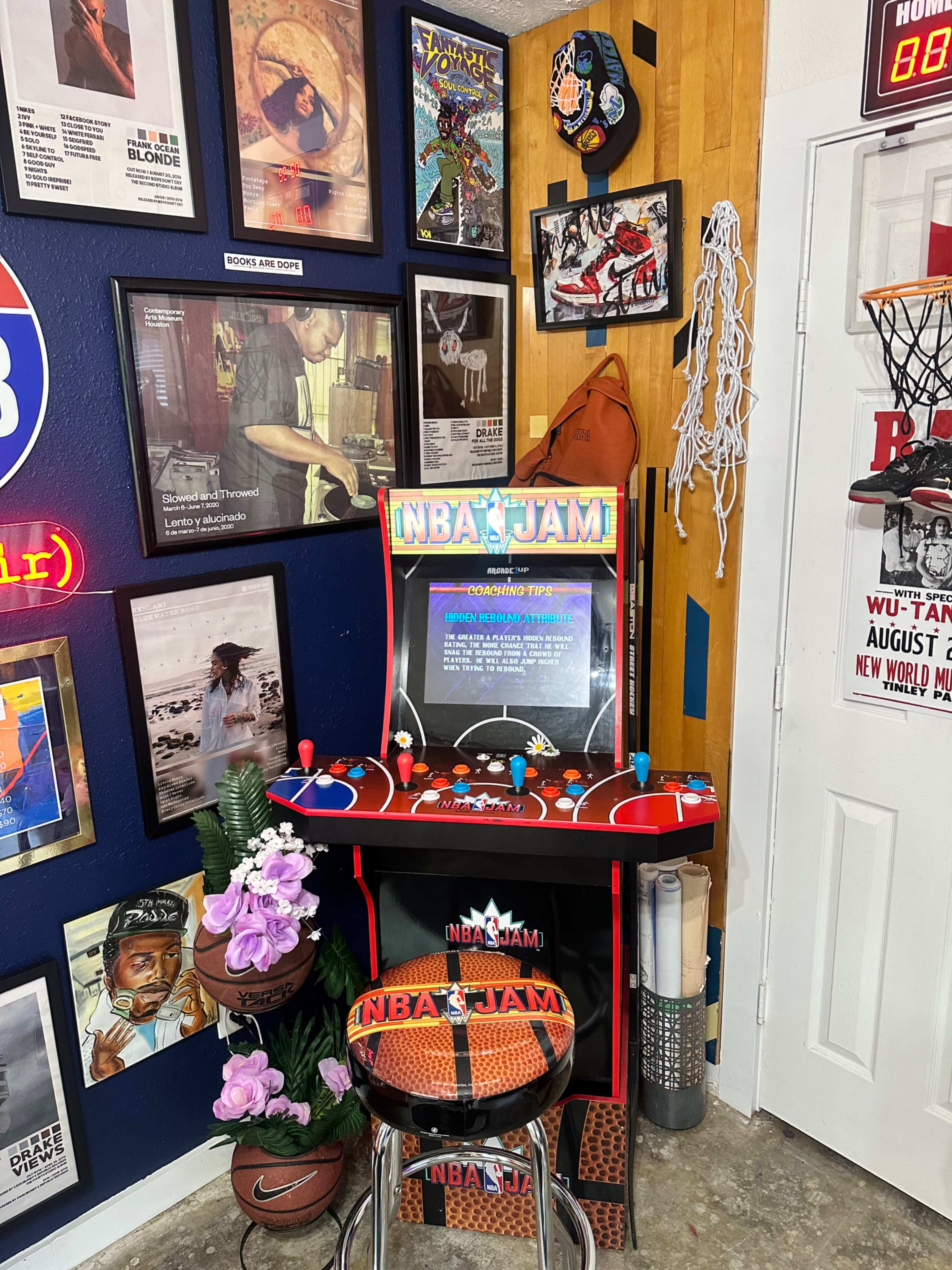 The image shows a vintage NBA Jam arcade game station surrounded by framed posters and memorabilia on a blue wall.