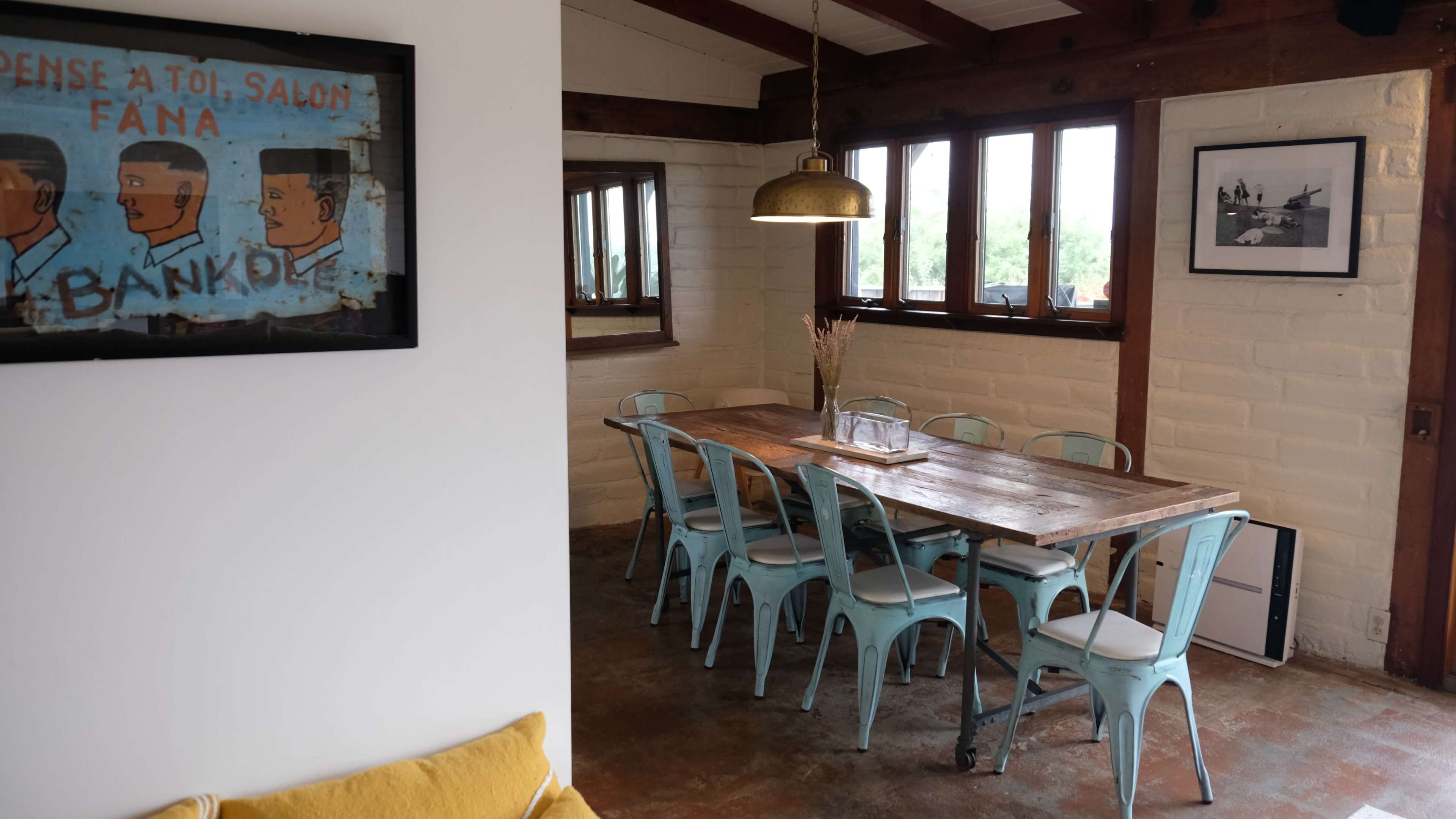 A rustic dining area featuring a wooden table surrounded by blue metal chairs, with large windows and a pendant light overhead.