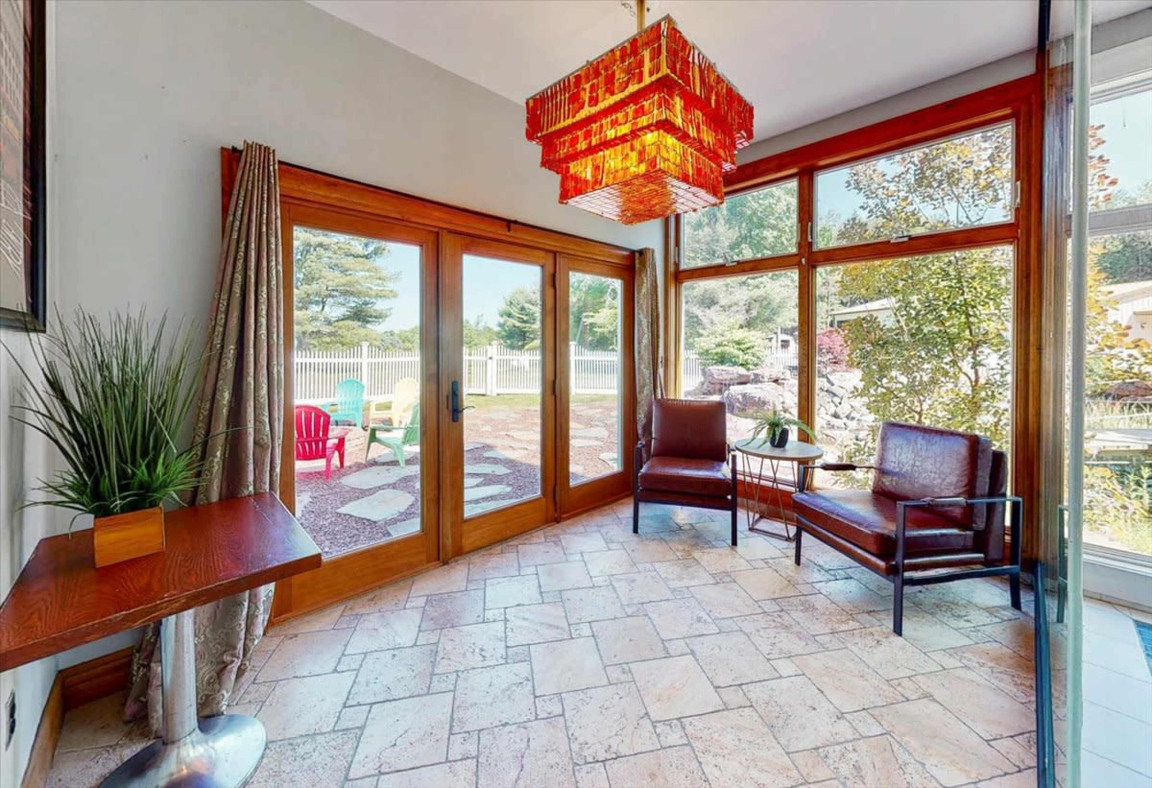 A sunroom with large glass doors, a decorative chandelier, and two chairs, overlooking a garden area with colorful outdoor furniture.
