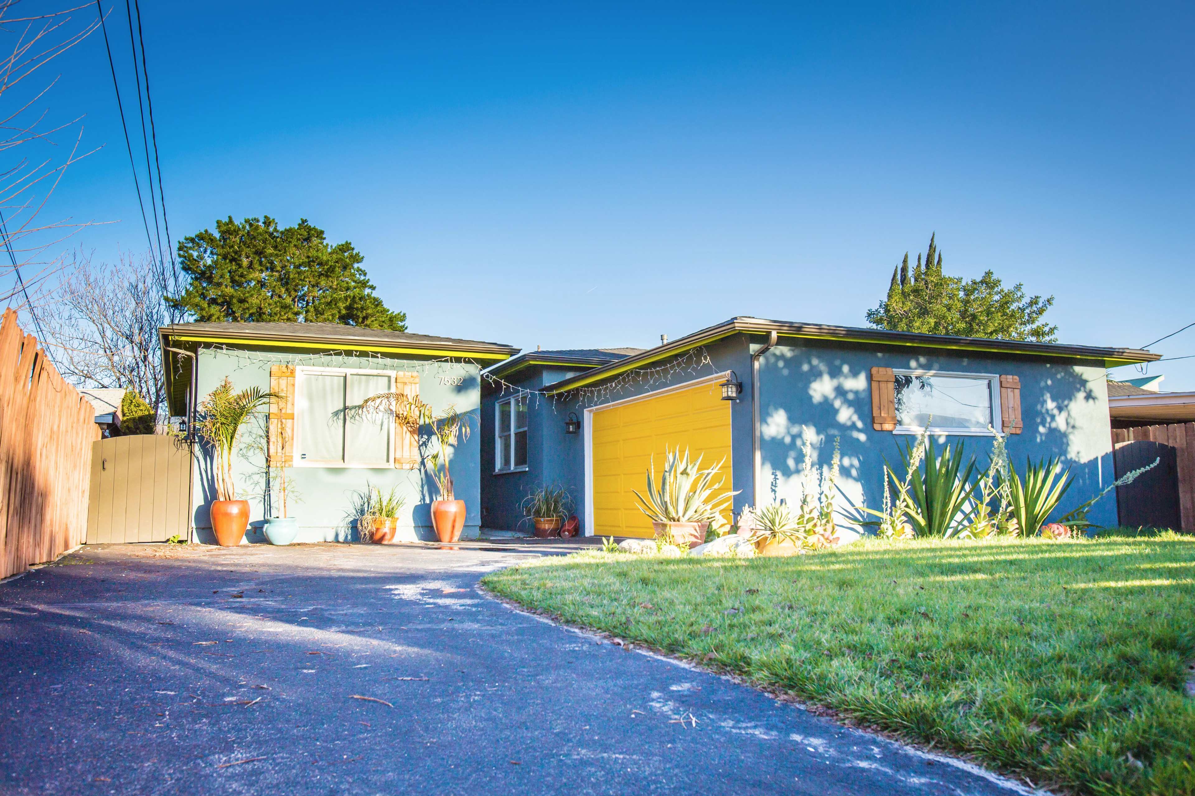 A blue house with a yellow garage door, surrounded by potted plants and a driveway leading up to it.