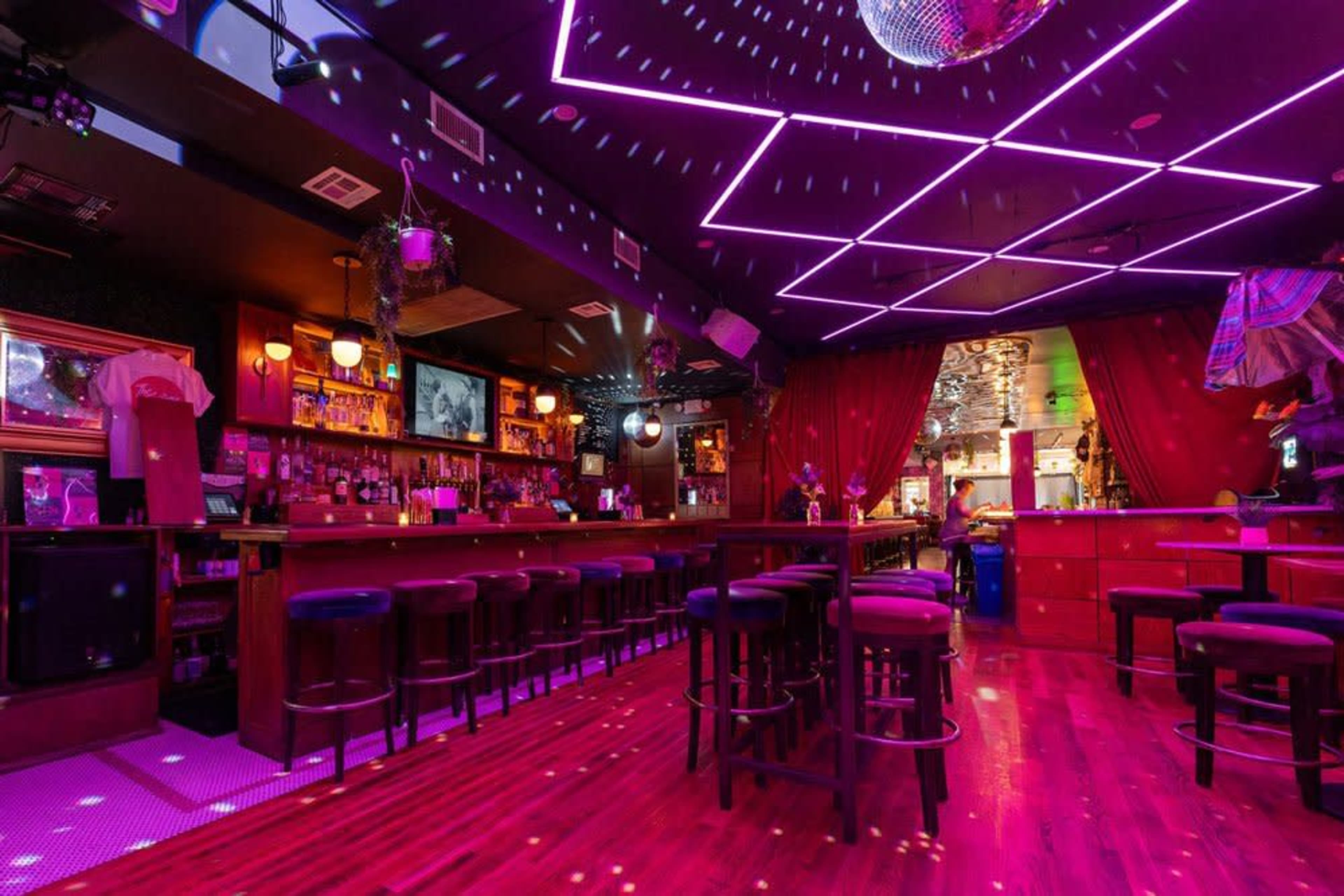 A vibrant bar interior features a long counter, illuminated by purple ceiling lights and a disco ball reflecting patterns across the room.