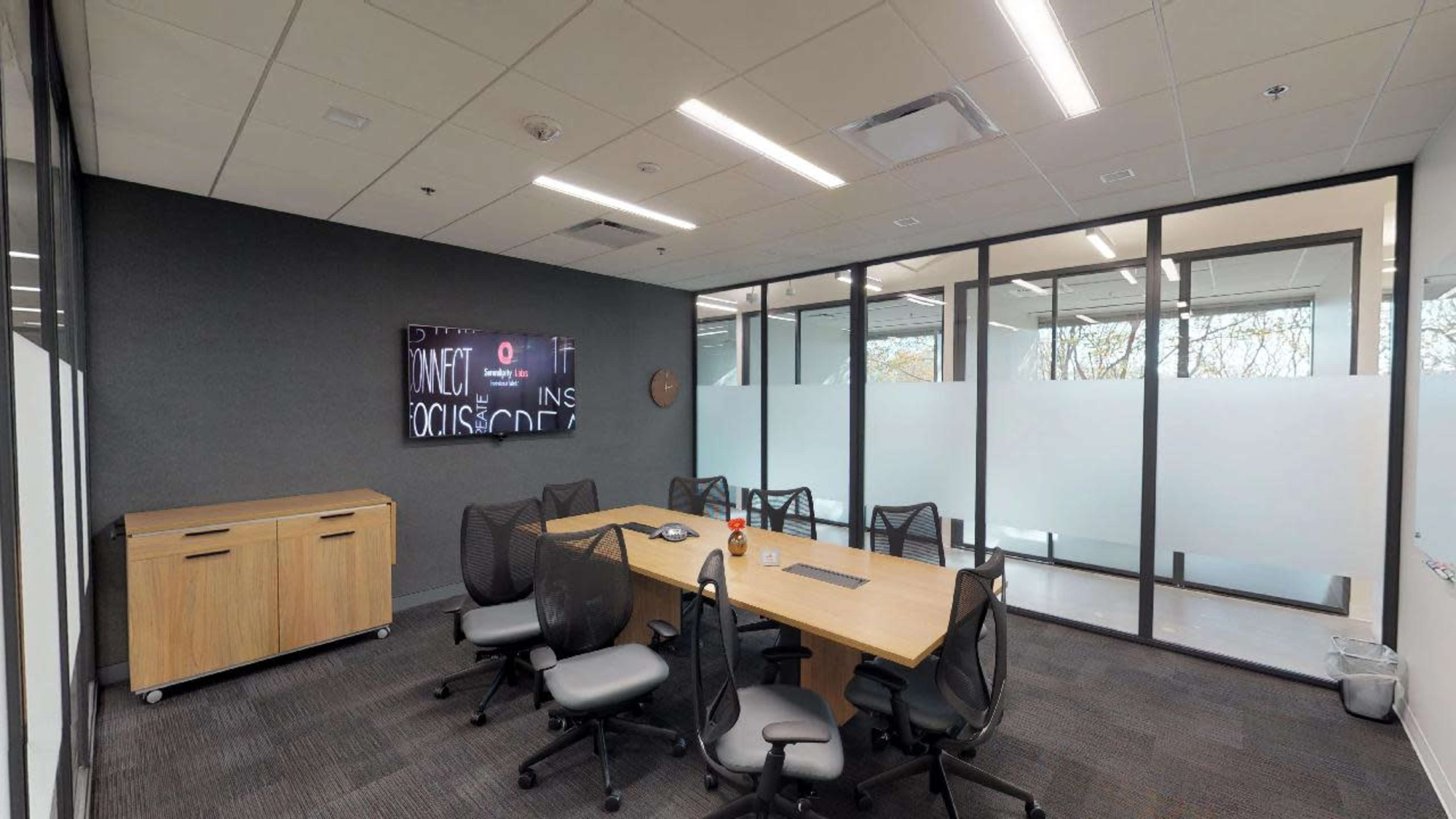 A modern conference room features a large wooden table surrounded by ergonomic chairs, with a wall-mounted screen and glass partitions.