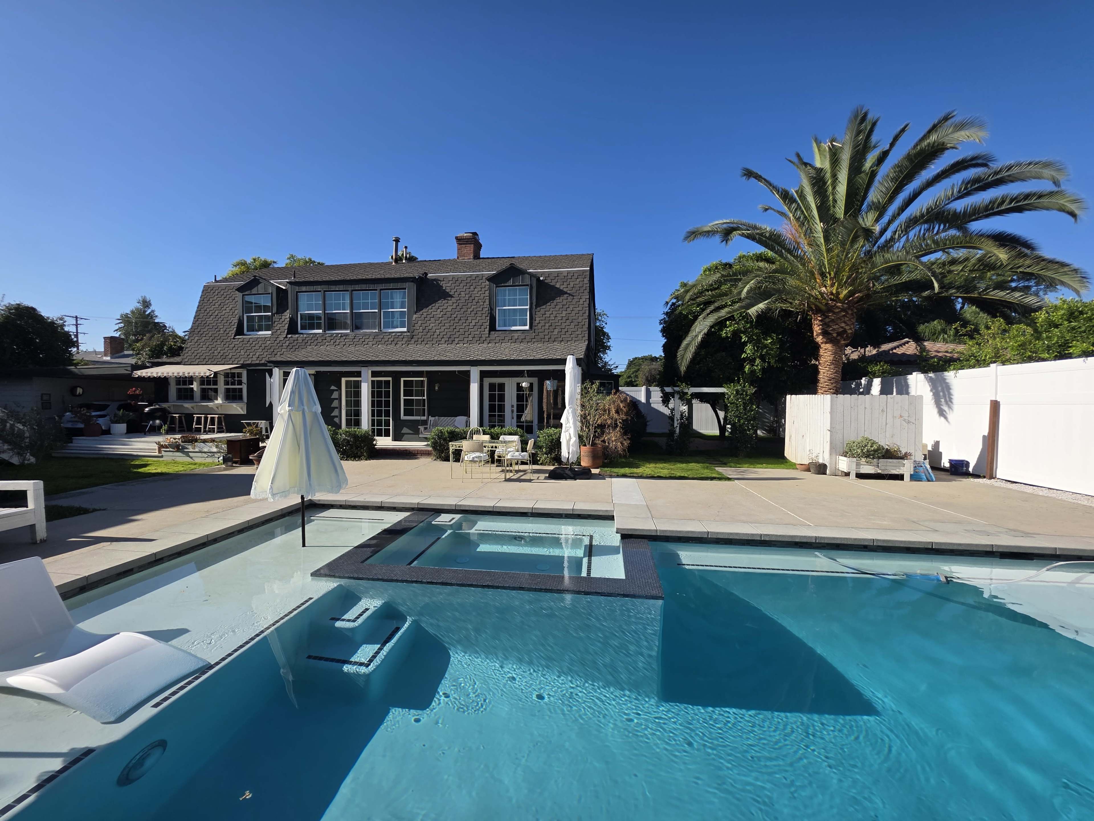 The image shows a backyard with a swimming pool and a two-story house surrounded by palm trees and outdoor seating areas.