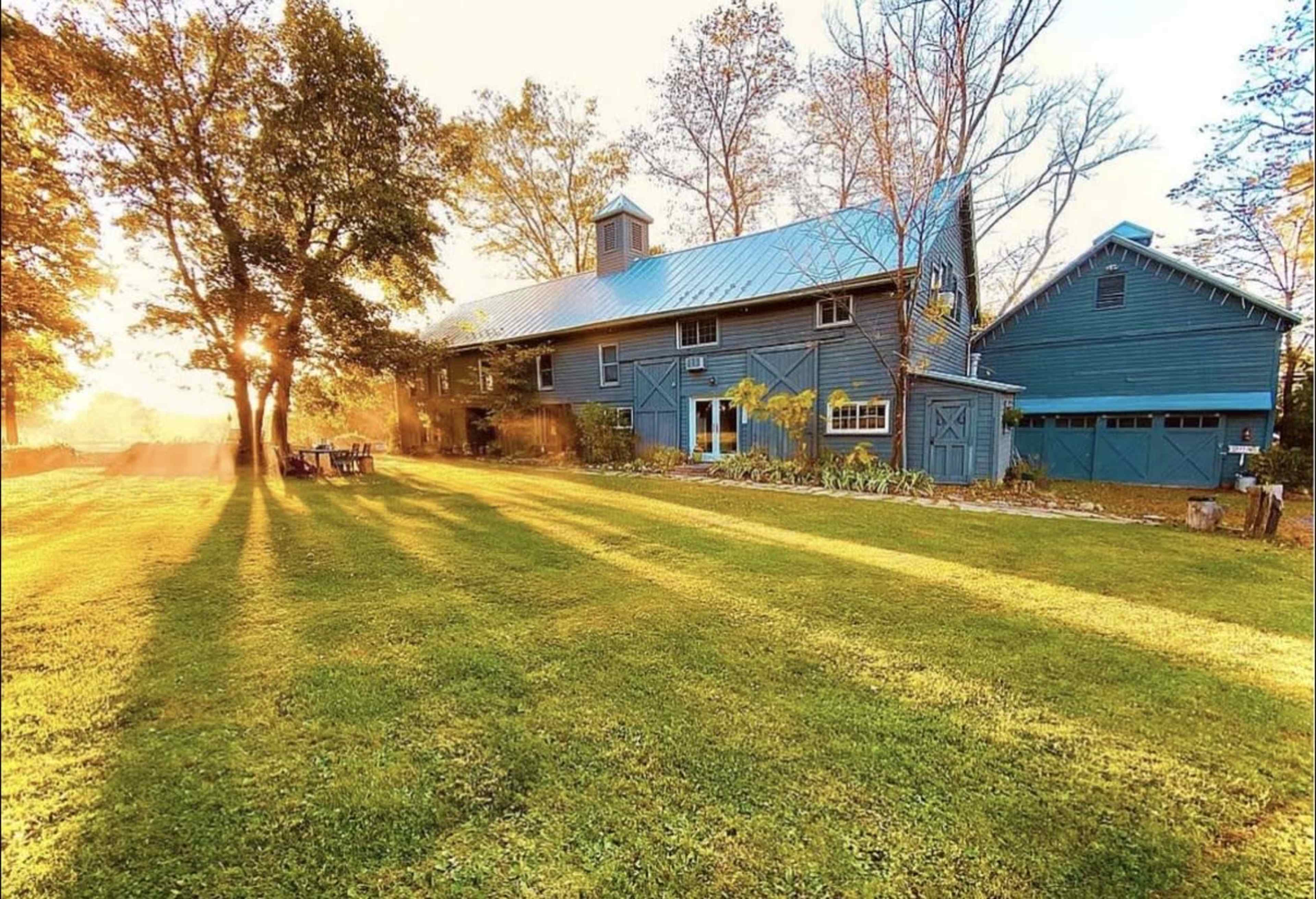 The image shows a large, blue barn-style house with a metal roof surrounded by green grass and trees, illuminated by warm light from a sunset.