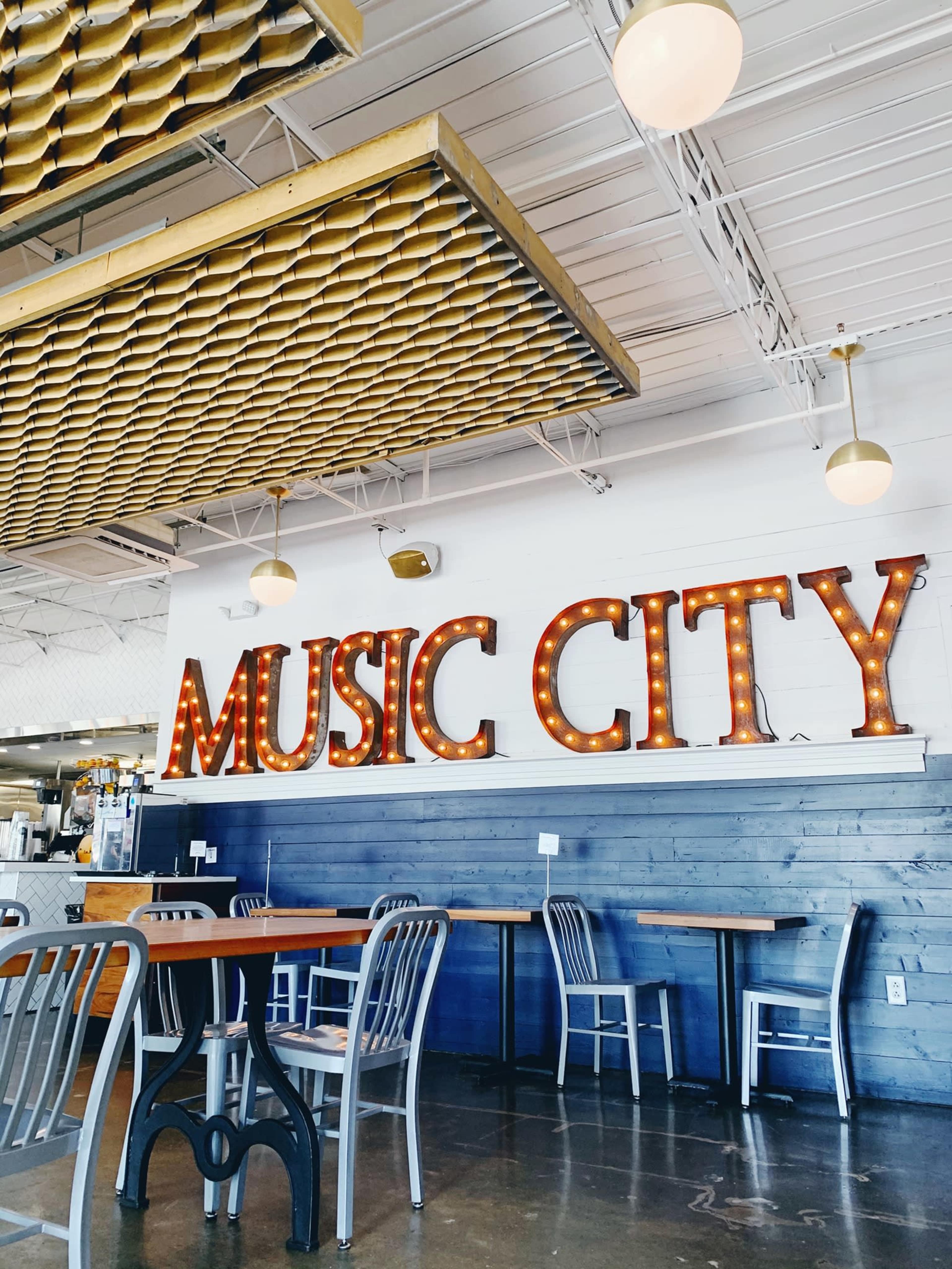 The image shows a brightly lit interior of a café or restaurant featuring a large, illuminated sign that reads "MUSIC CITY" on one wall, with wooden tables and metal chairs arranged in the space.