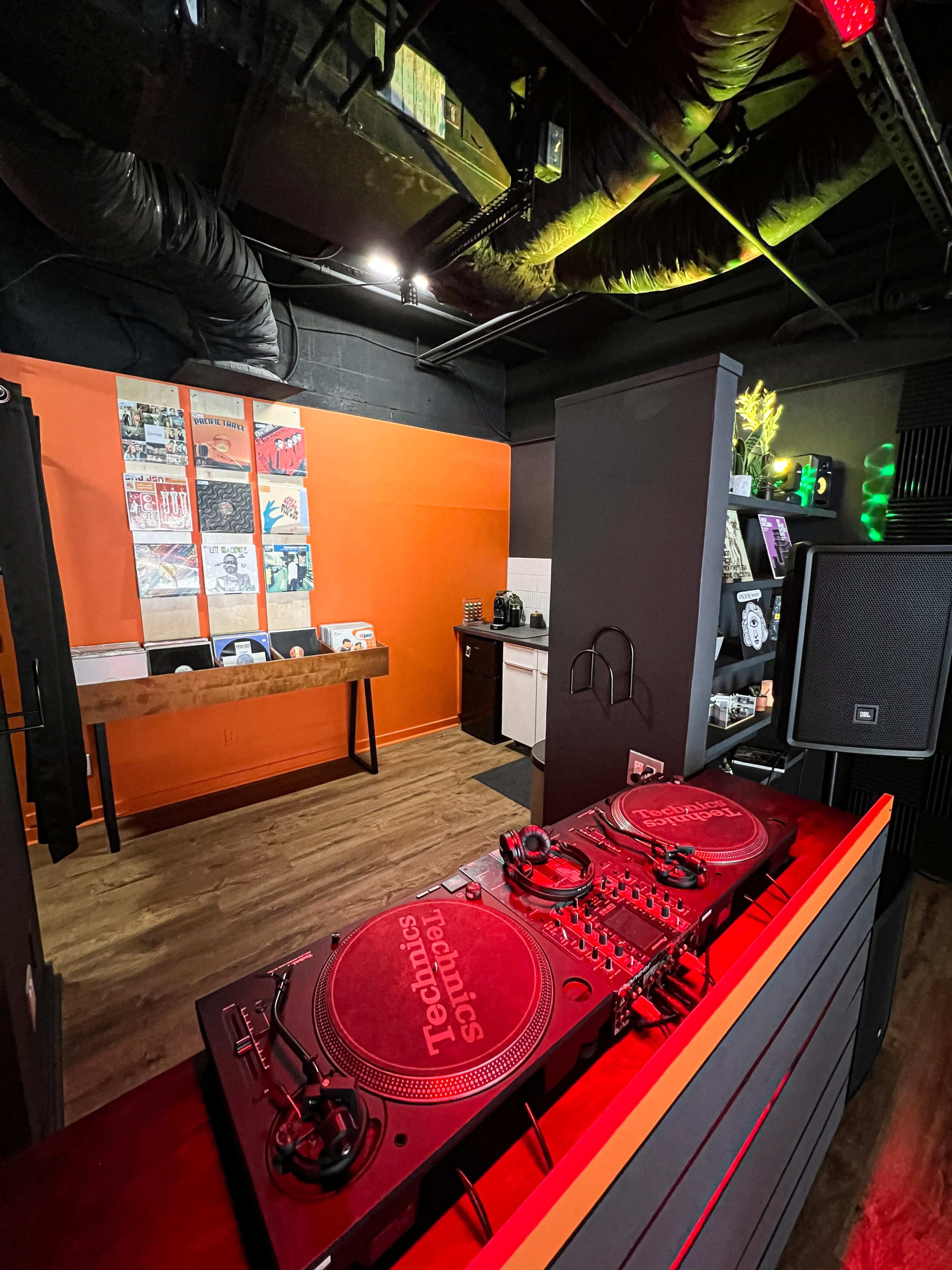 A music studio featuring a DJ setup with turntables in the foreground, a kitchen area in the background, and walls decorated with artwork.