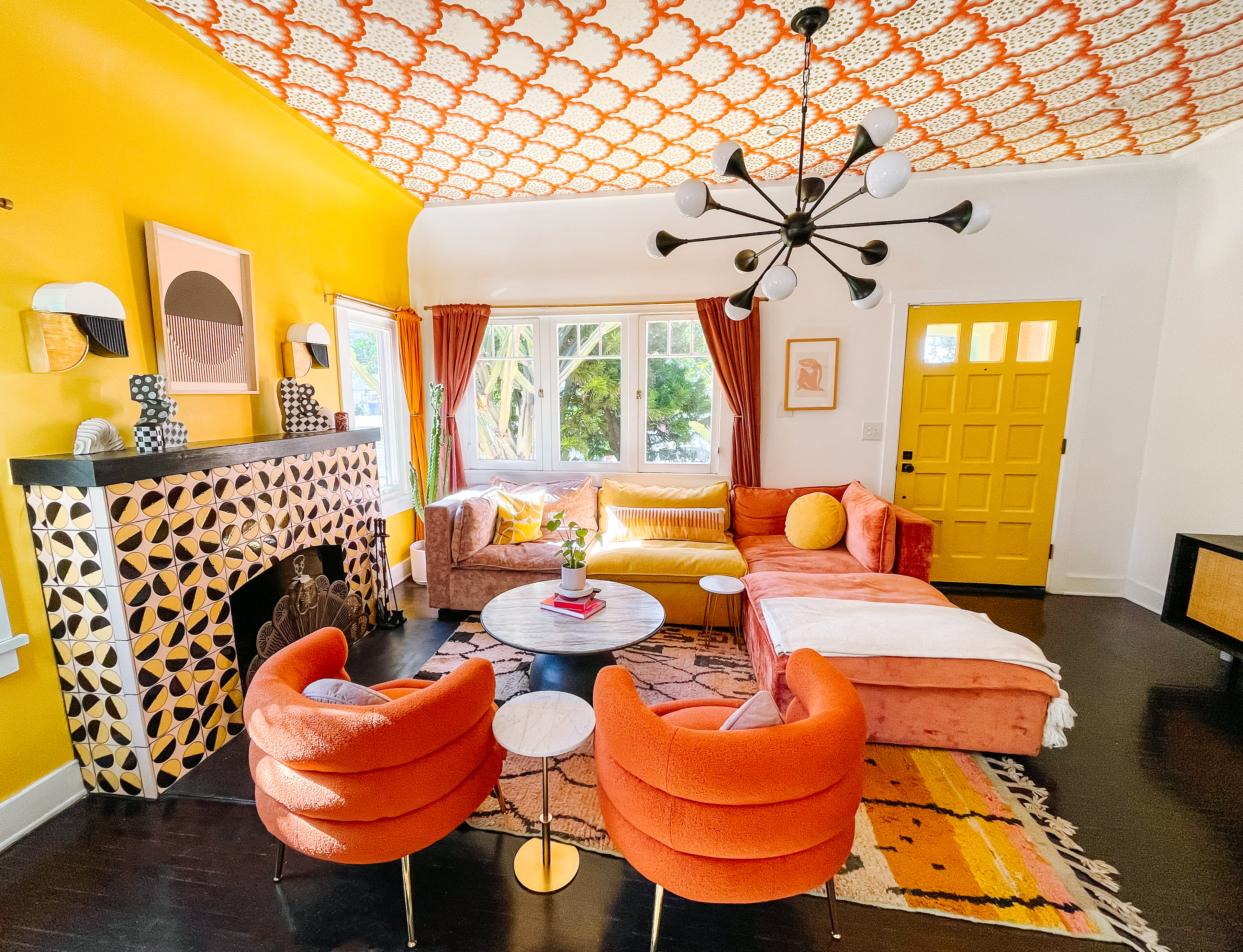 The living room features a bright yellow wall, ornate patterned ceiling, and a colorful mix of furniture including an orange and pink sofa, paired with round chairs and a glass coffee table.