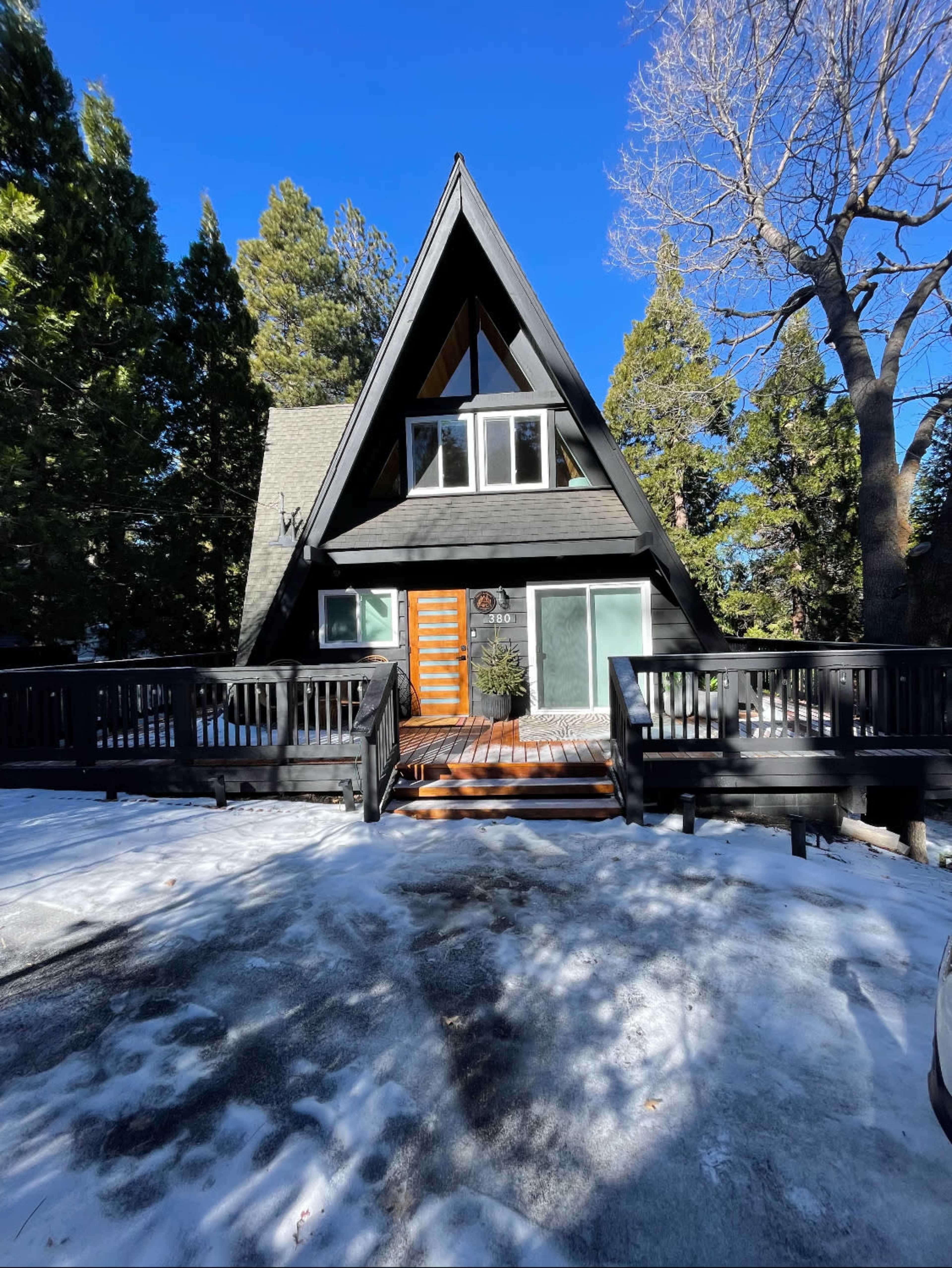 A black A-frame house with large windows and a wooden front door is set against a snowy landscape and surrounded by tall evergreen trees.