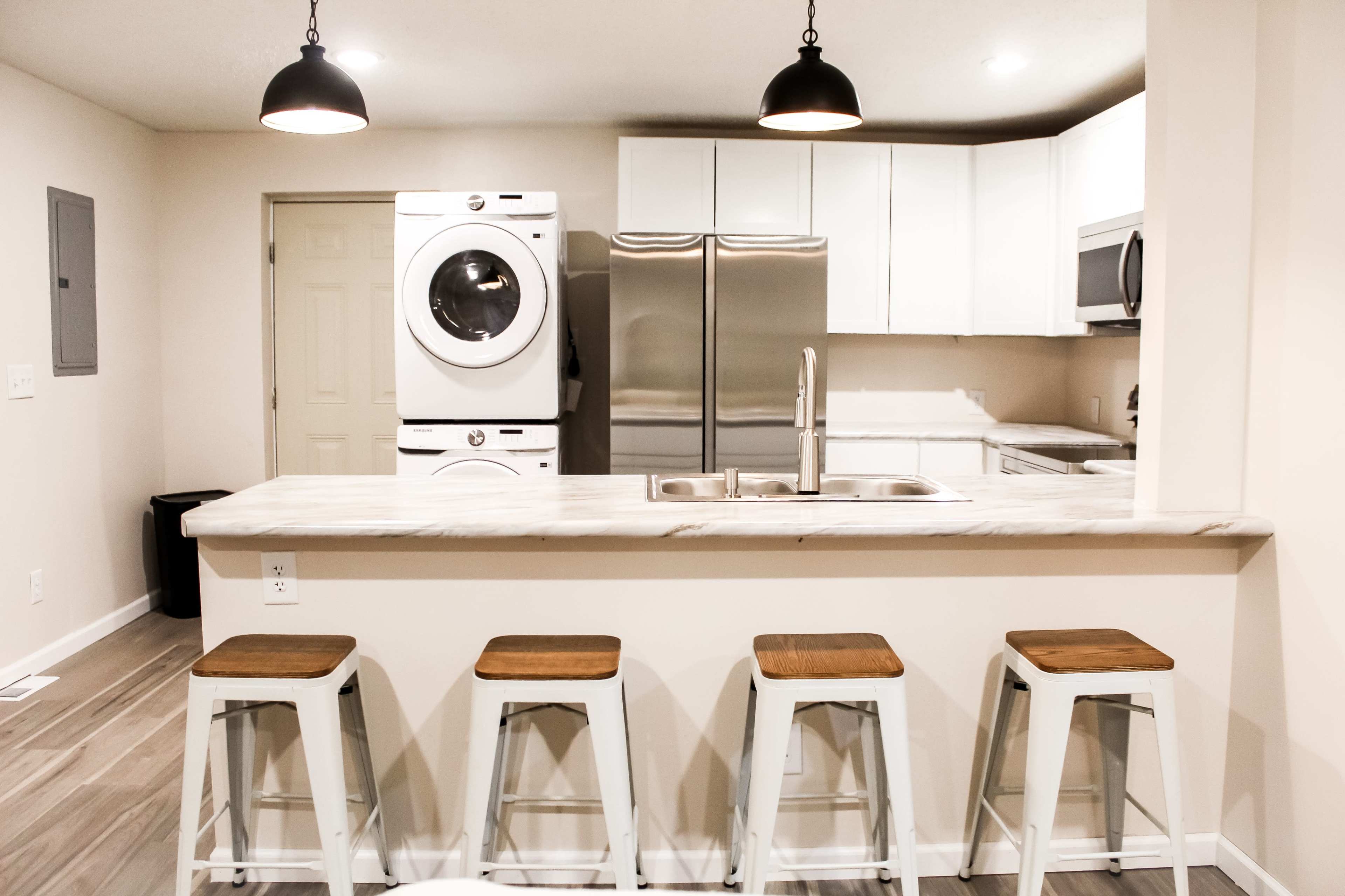 The image shows a modern kitchen with a washer and dryer unit, stainless steel appliances, and four metal bar stools at a marble countertop.