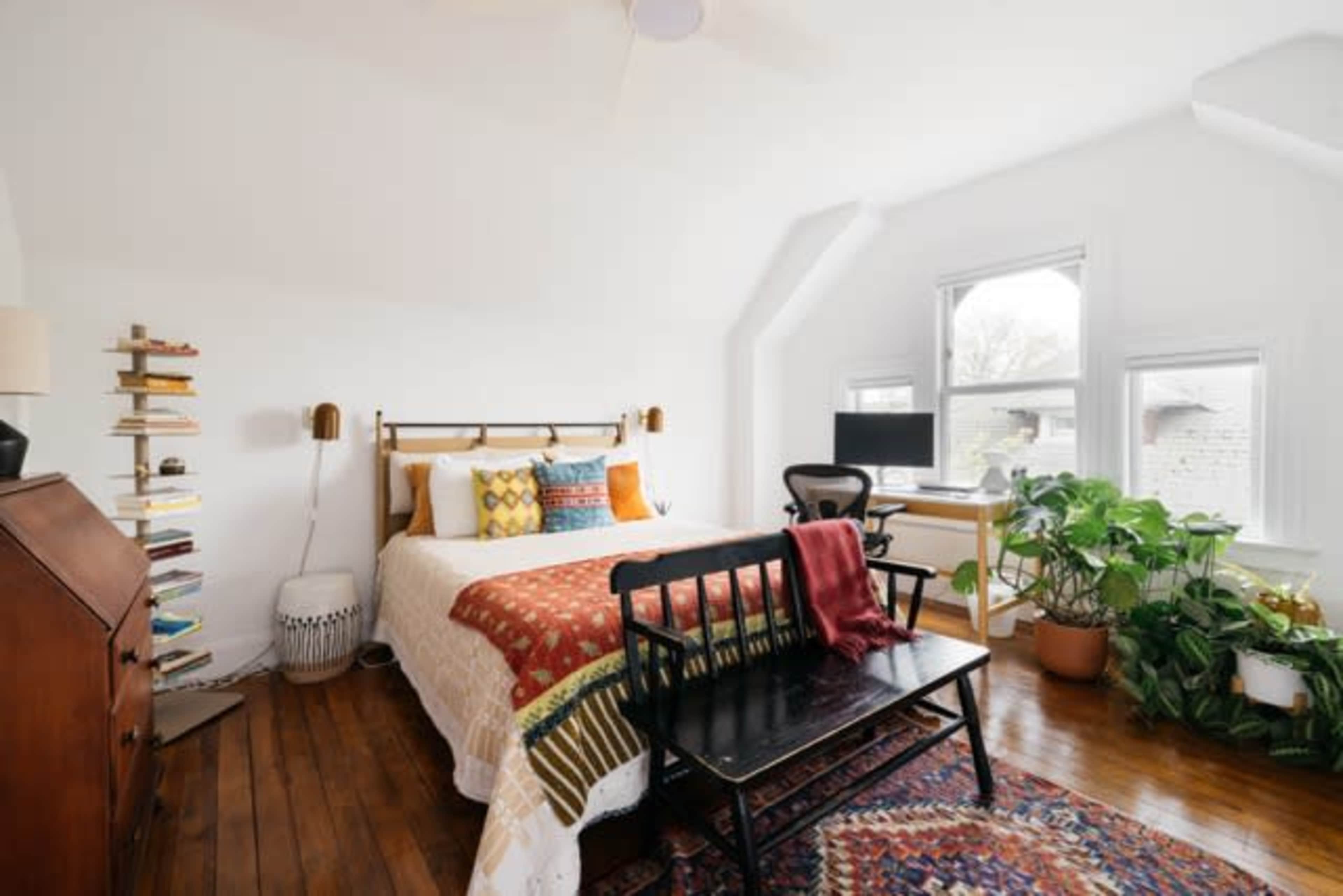 A cozy bedroom features a bed with colorful pillows, a desk with a computer, and a potted plant by the window.