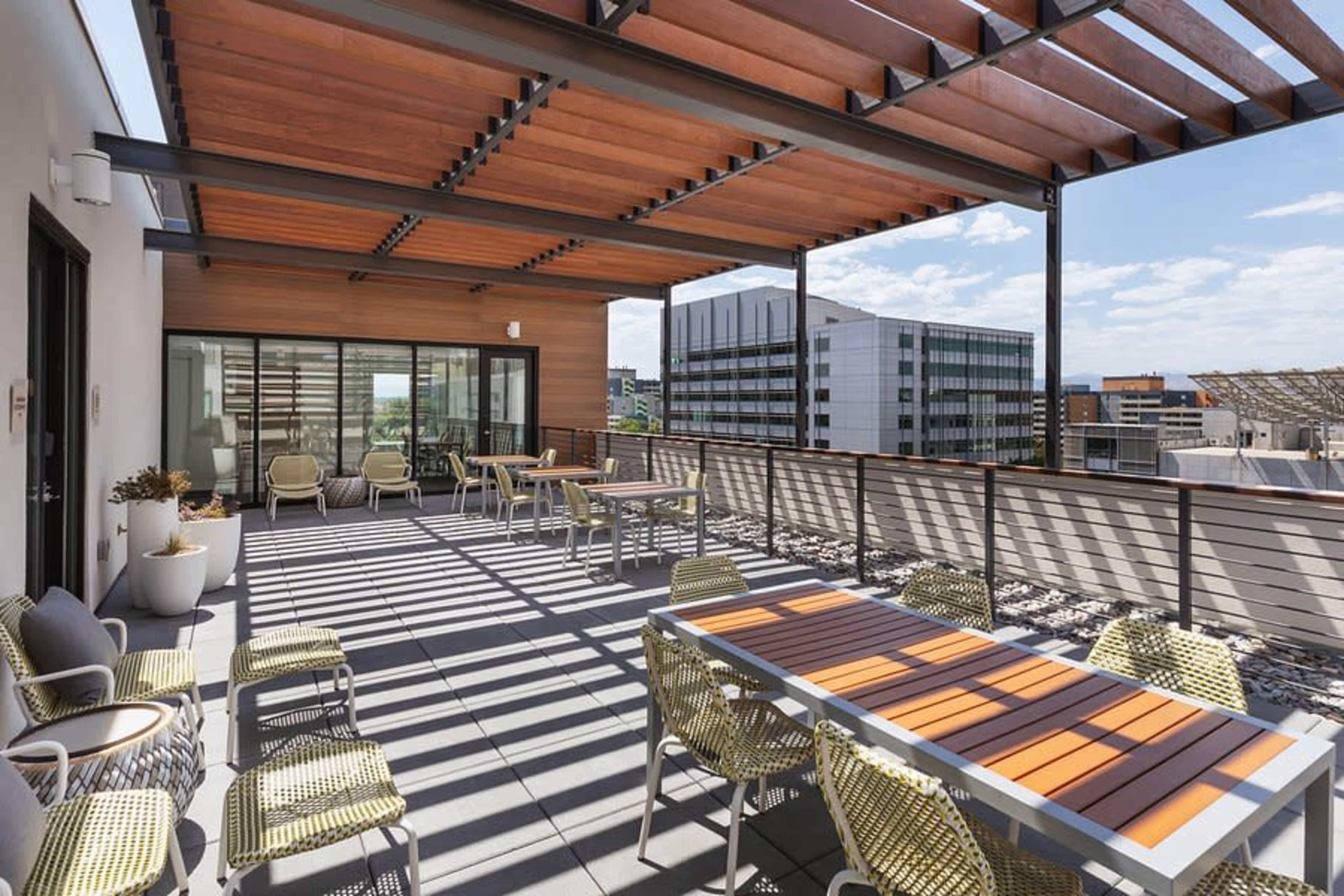 A modern rooftop terrace features seating areas and tables under a wooden pergola, with a view of surrounding buildings and a clear blue sky.