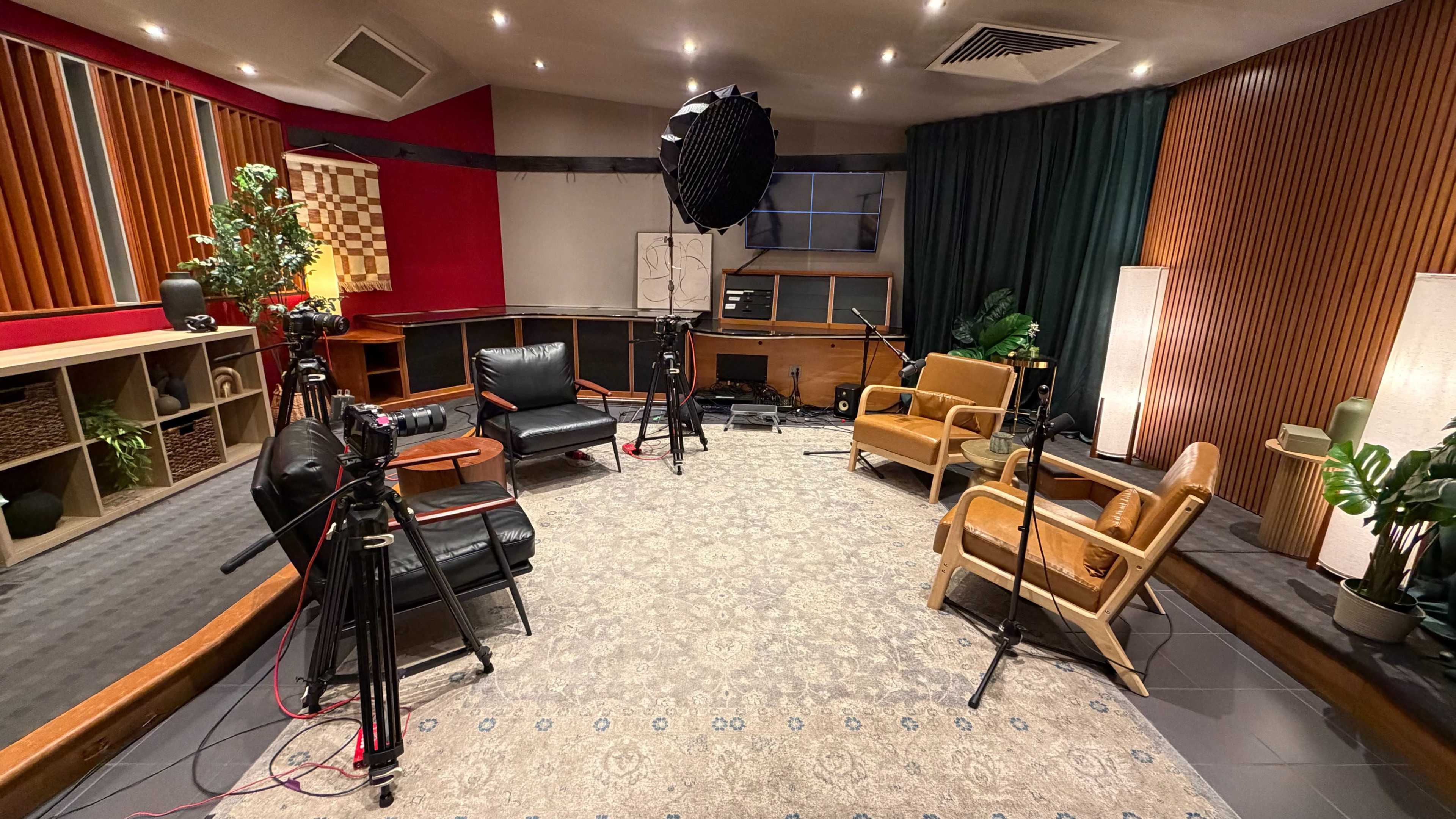 The image shows a spacious recording studio with black and brown chairs, three cameras on tripods, and plants arranged around a large patterned rug.