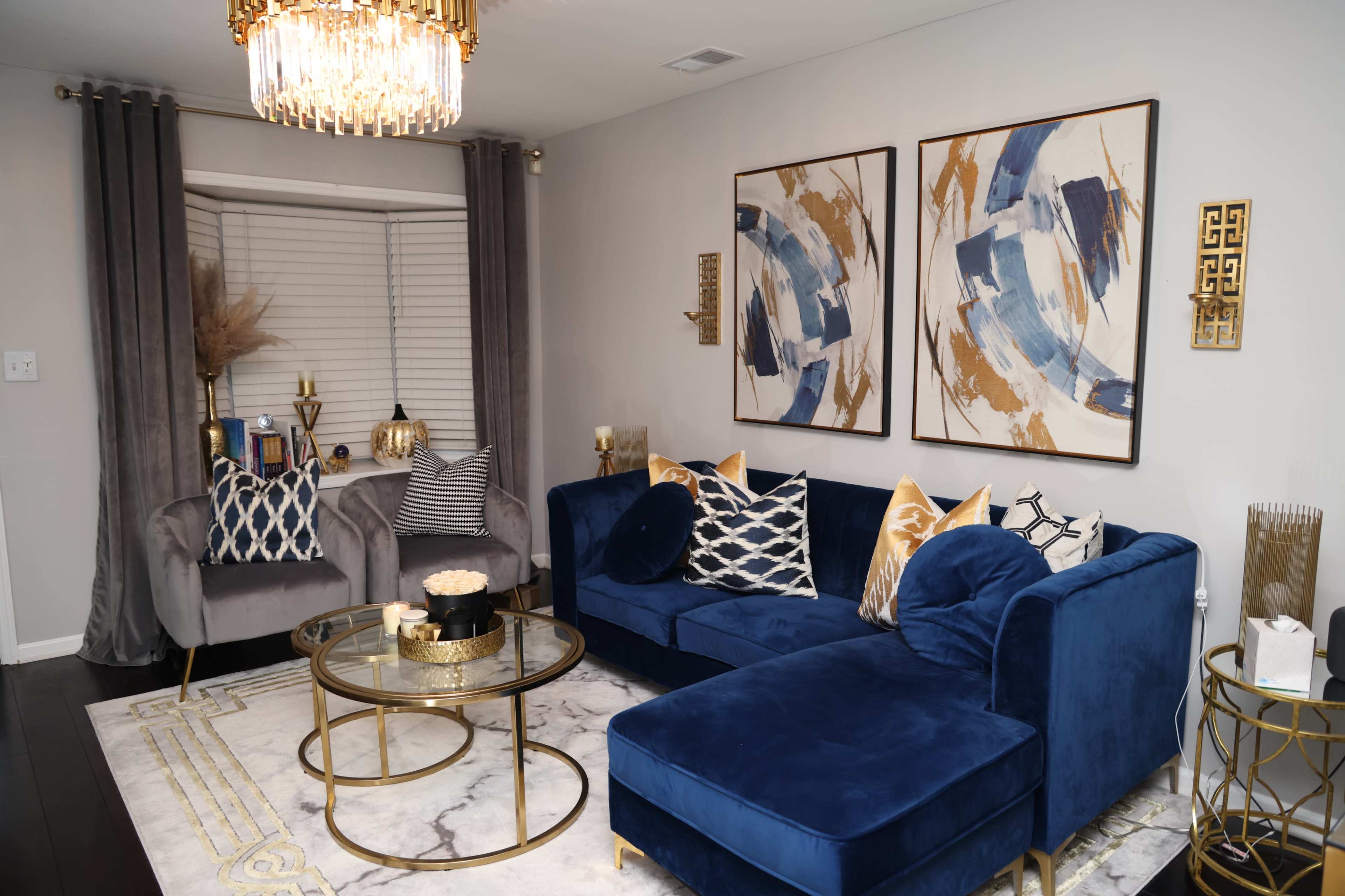 A modern living room featuring a blue sofa, a gray armchair, circular gold tables, and abstract wall art above a marble-patterned rug.