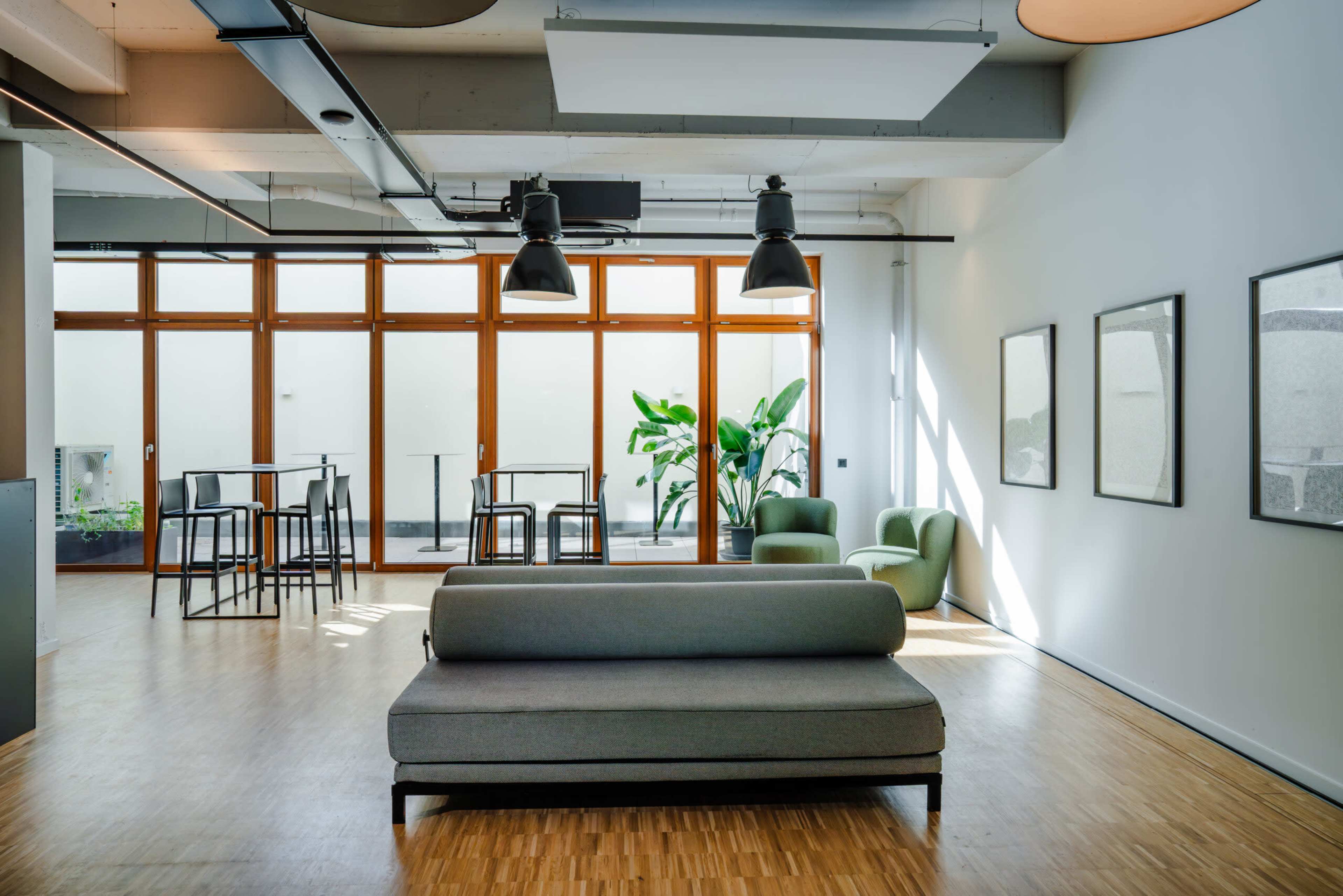 The image shows a modern, minimalistic lounge area featuring a gray sofa, two green chairs, and a table with high stools, illuminated by large windows that allow natural light to enter.