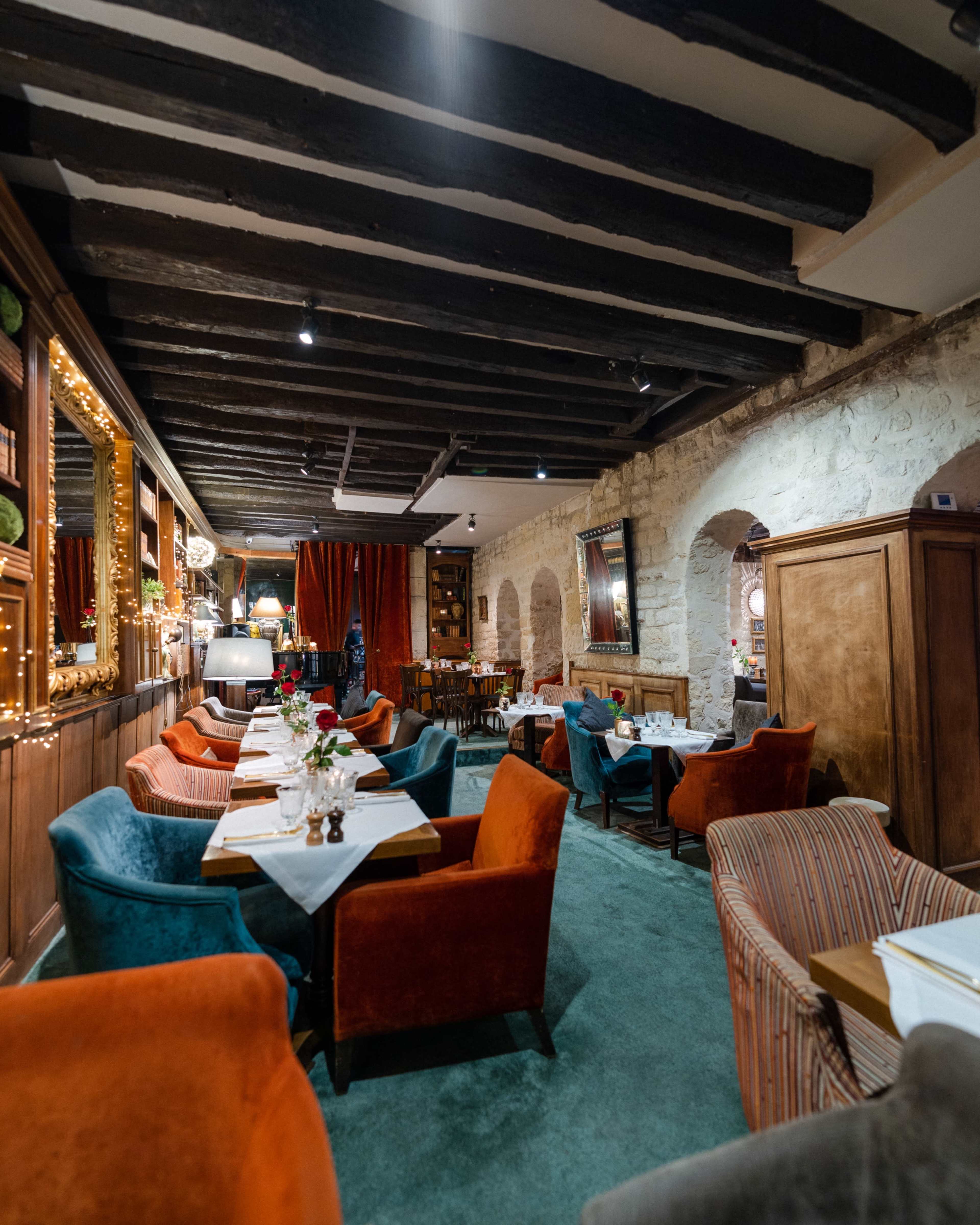 The image shows a cozy restaurant interior featuring a mix of colorful upholstered chairs, wood-paneled walls, and warm lighting.