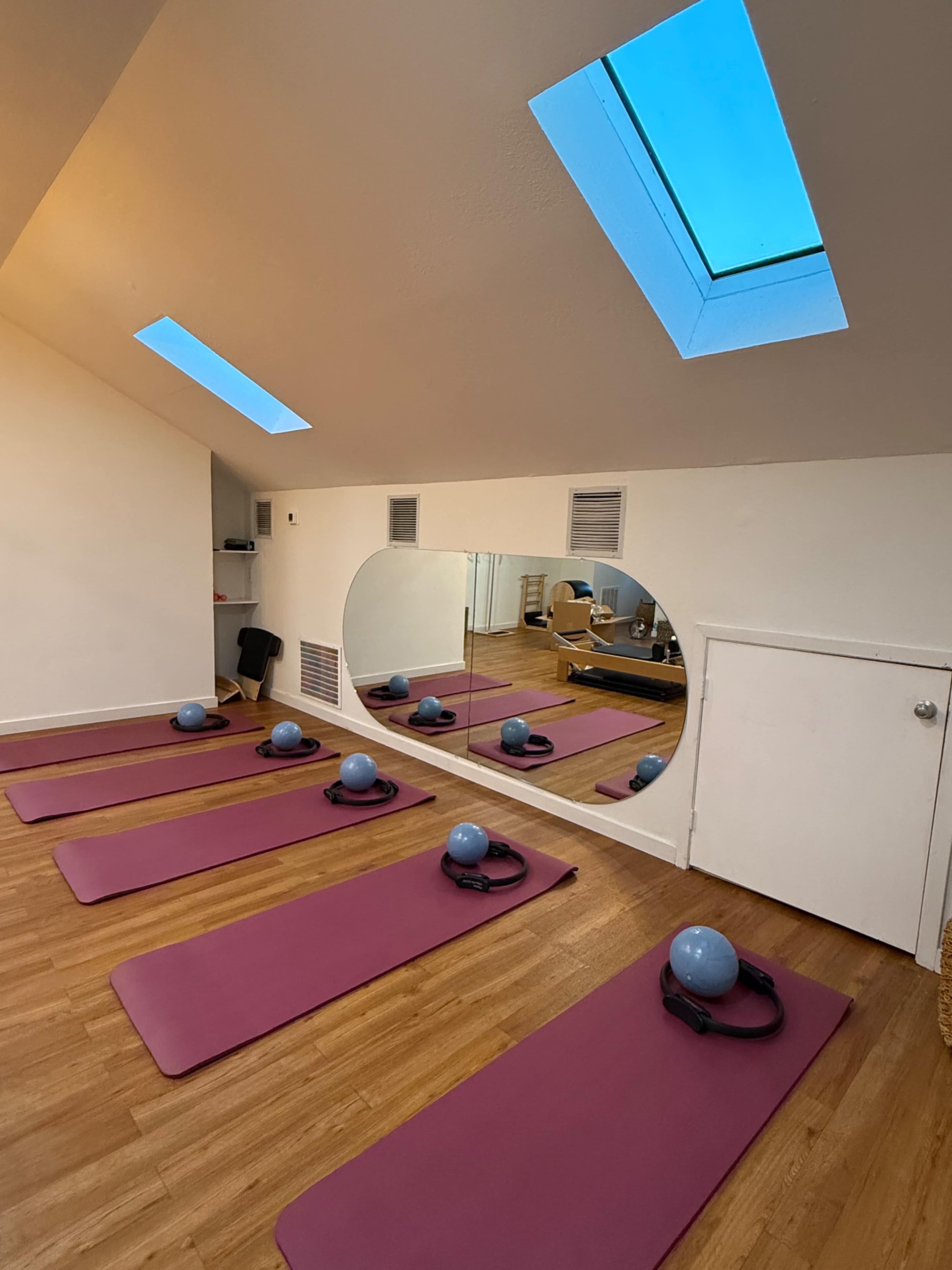 The image shows a Pilates studio with several exercise mats arranged on a wooden floor, blue stability balls on top, and a large mirror reflecting the skylights above.