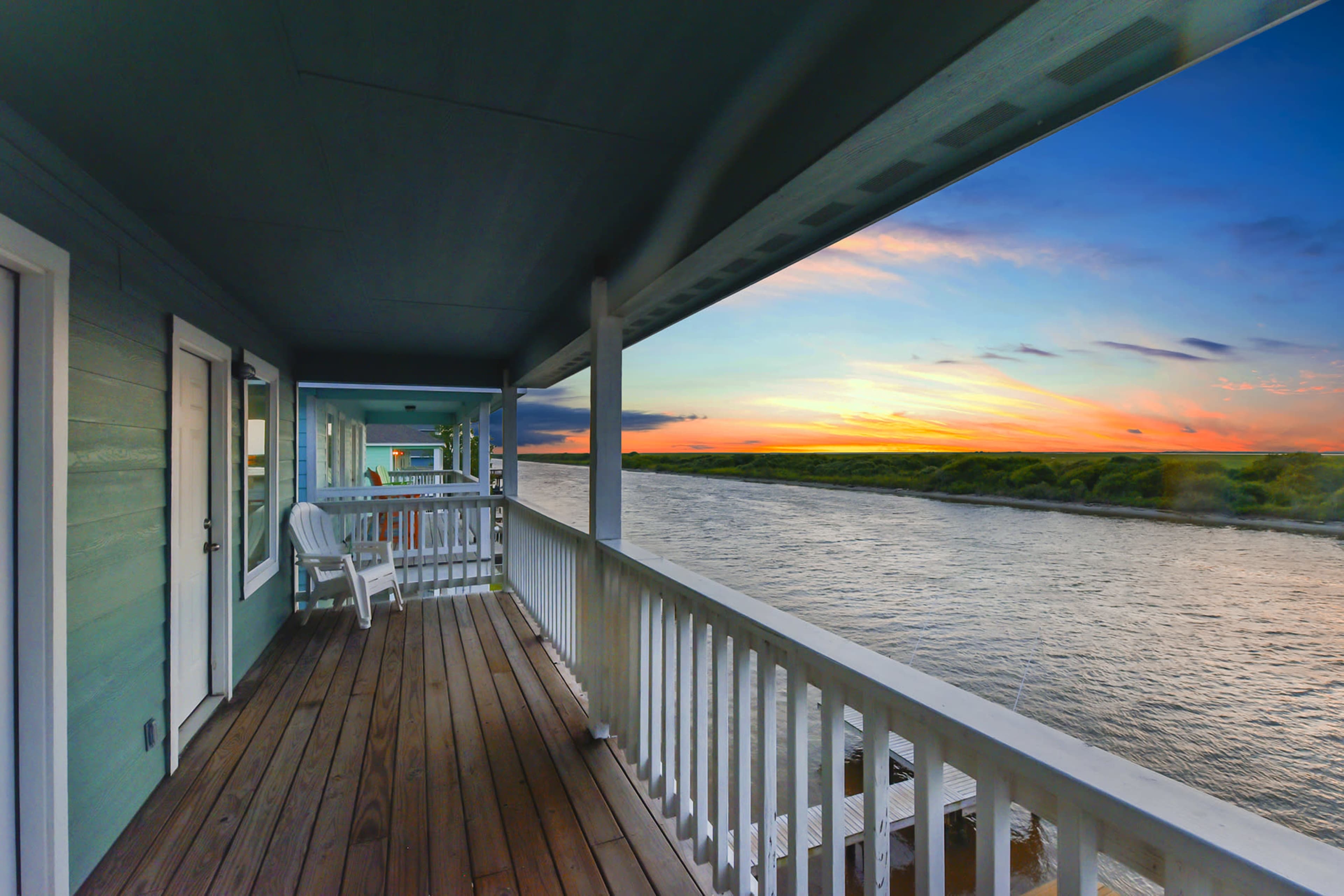 The image shows a wooden balcony with white railings overlooking a river at sunset.
