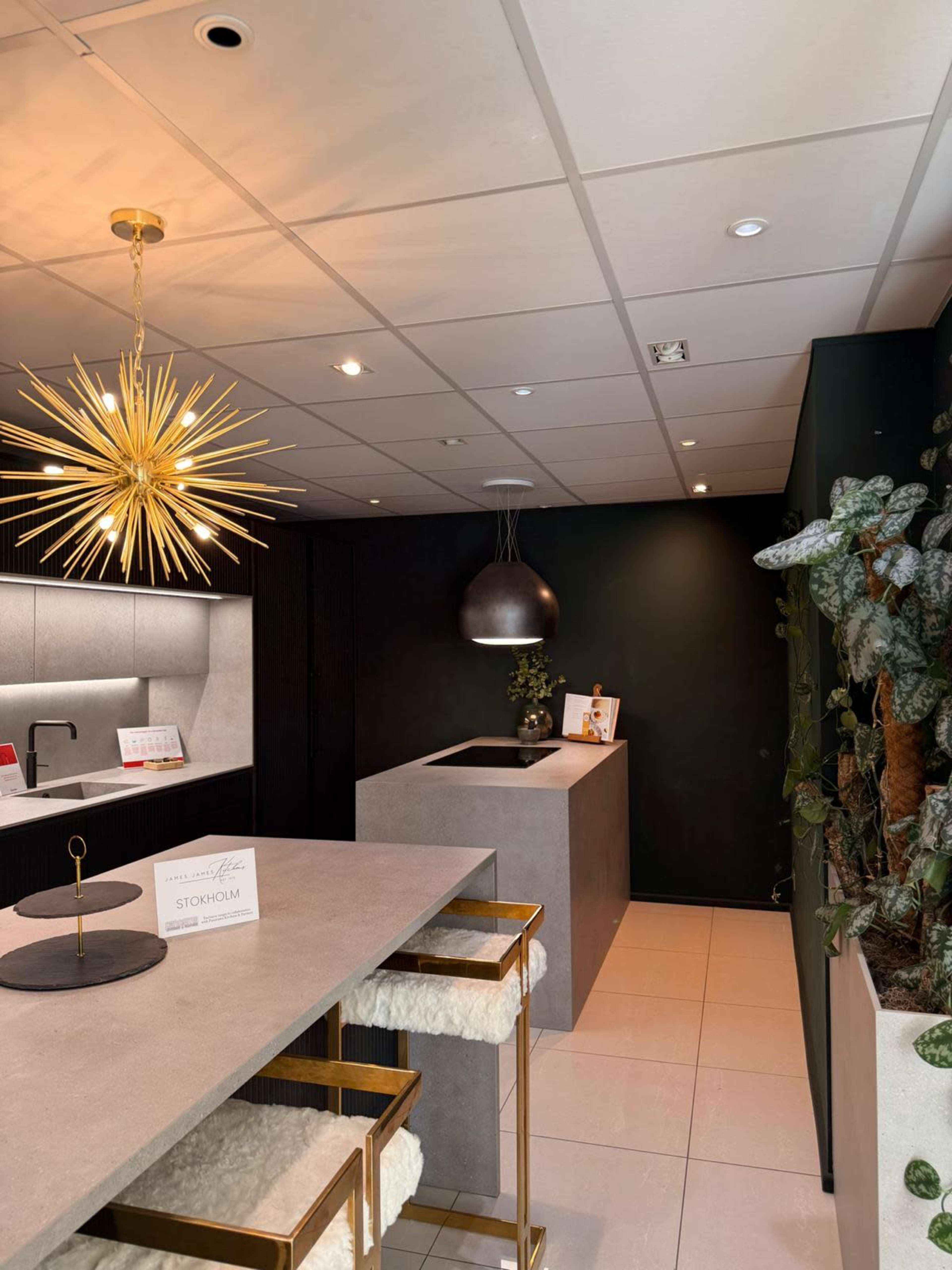 The image shows a modern kitchen or showroom with a sleek countertop, geometric light fixture, and potted plants in a contemporary setting.