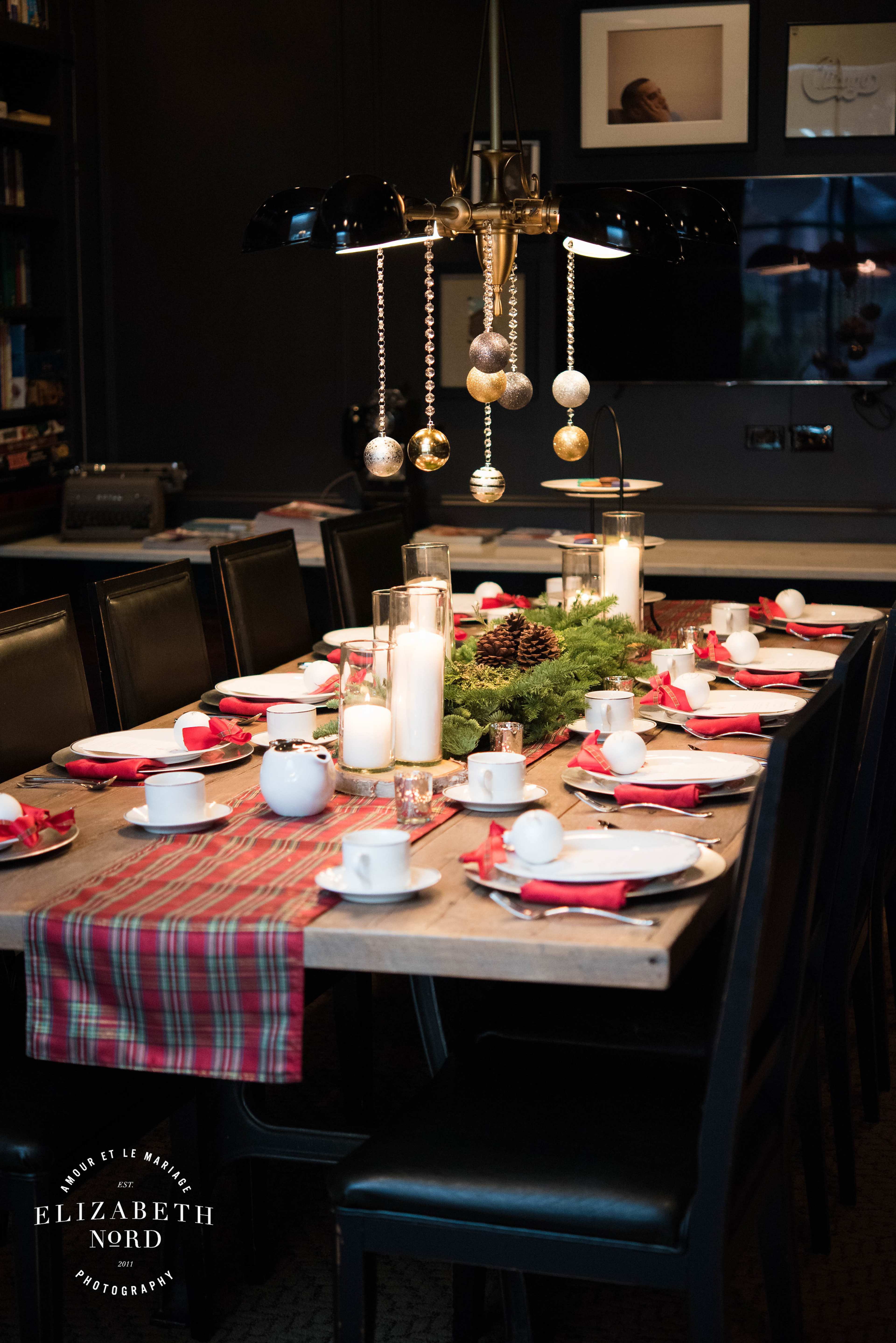 A dining table is set with white dishes, red napkins, and decorative centerpieces, illuminated by a chandelier adorned with ornaments.