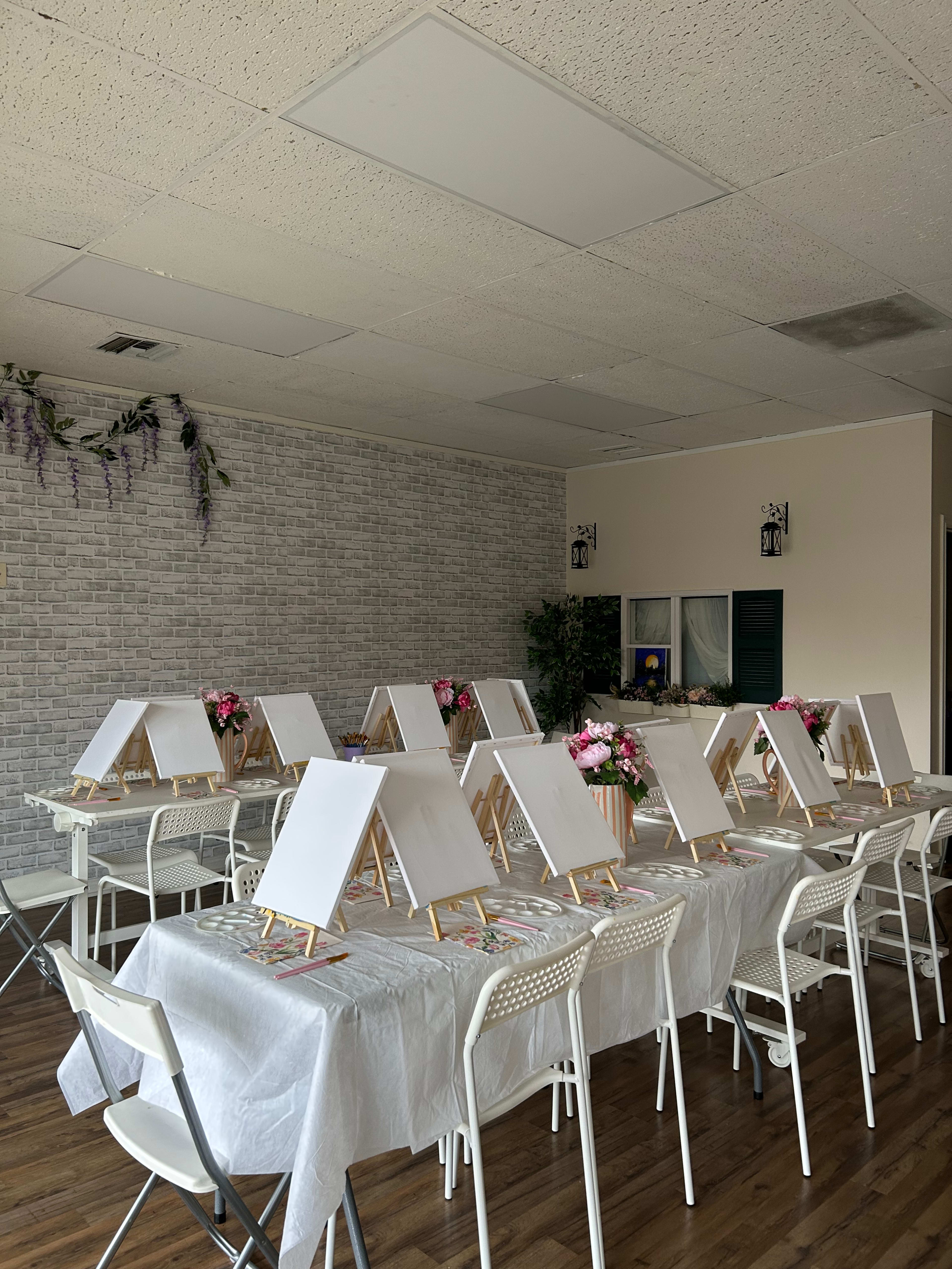 The image shows an art studio setup with several empty easels on tables adorned with white tablecloths and floral centerpieces.