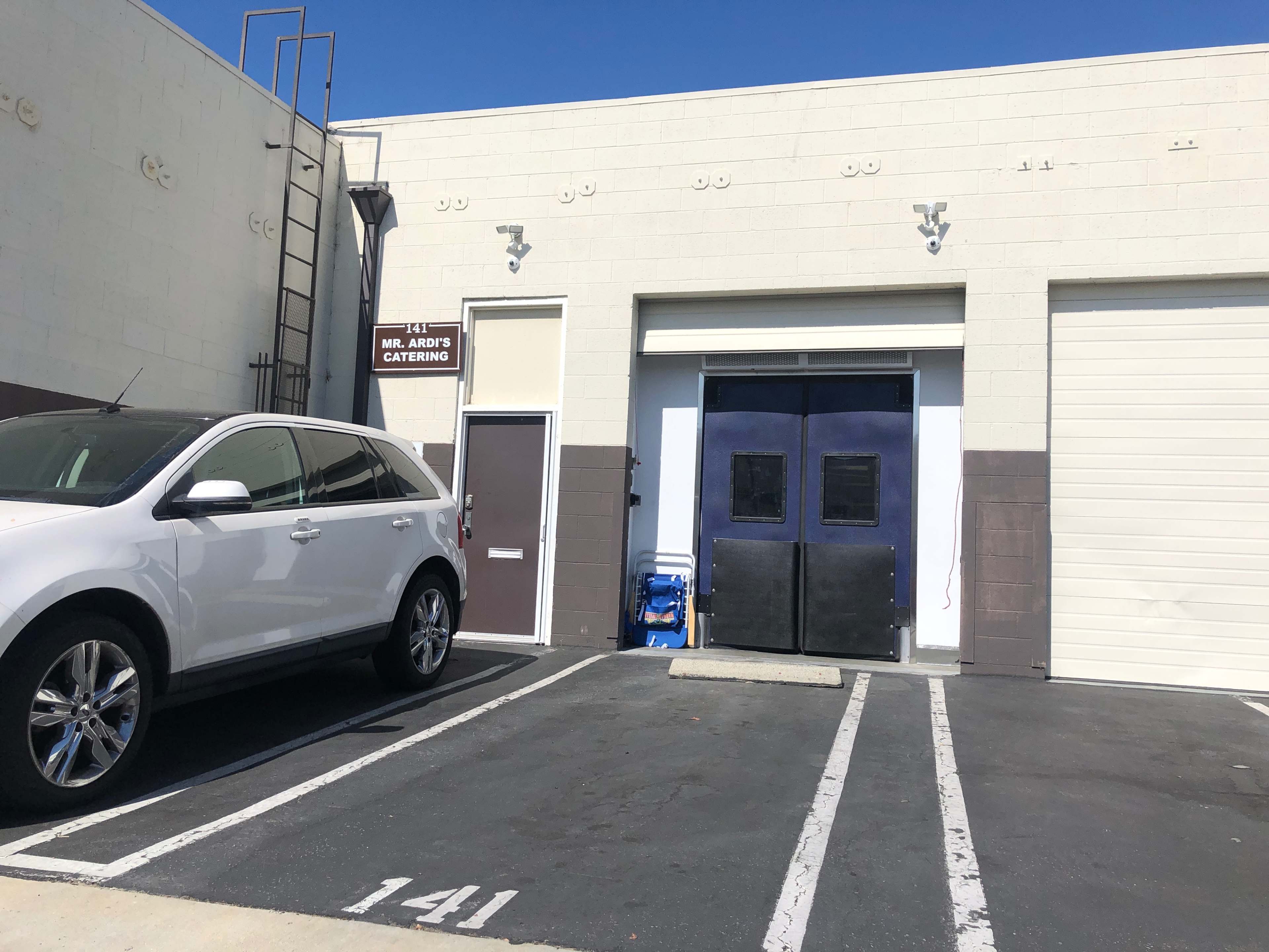 A white vehicle is parked in front of a building with two large doors, one blue and one white, marked for Mr. Arto's Catering.