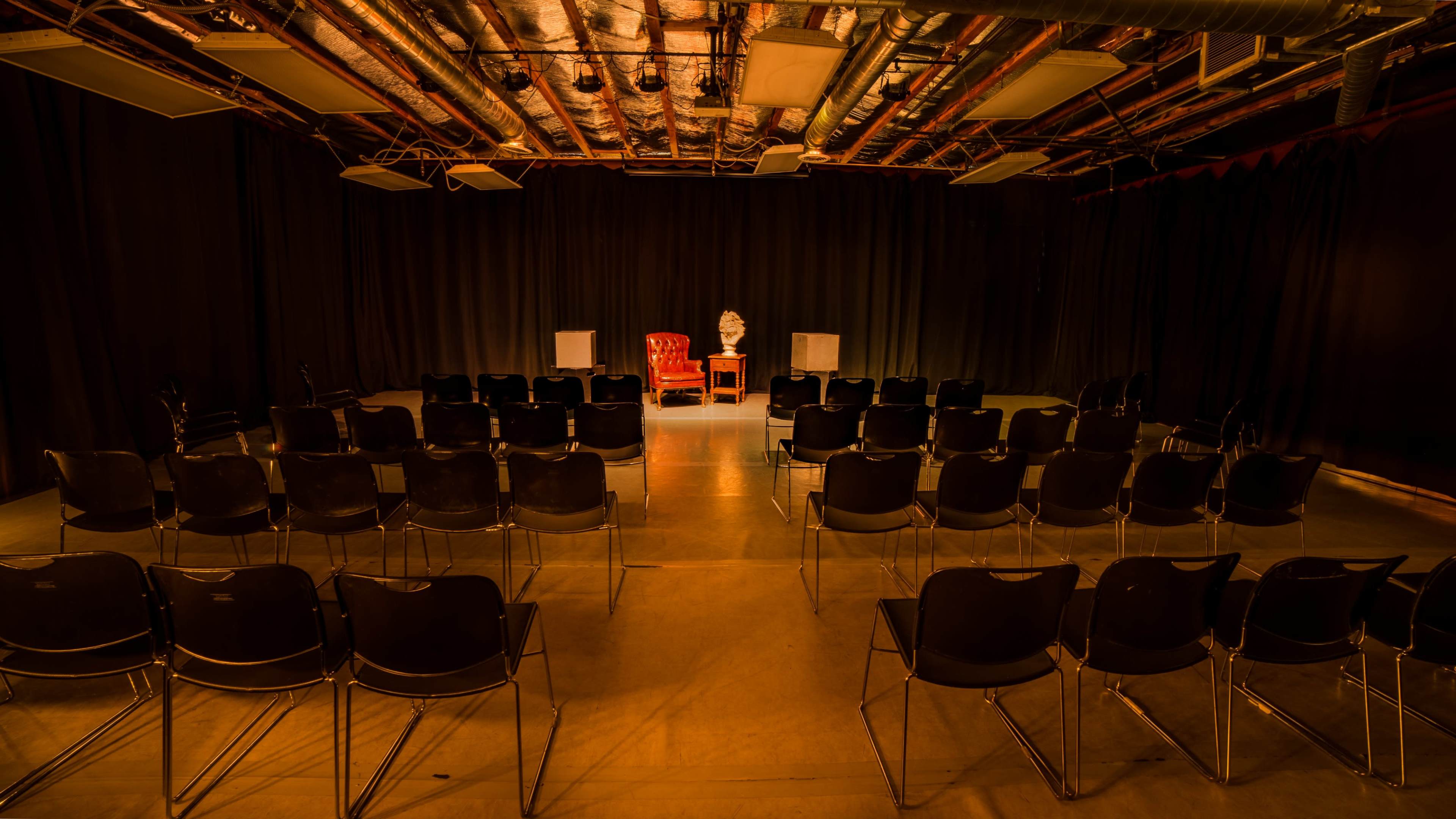 A red armchair is positioned on a stage in front of a row of chairs in a dimly lit theater setting.