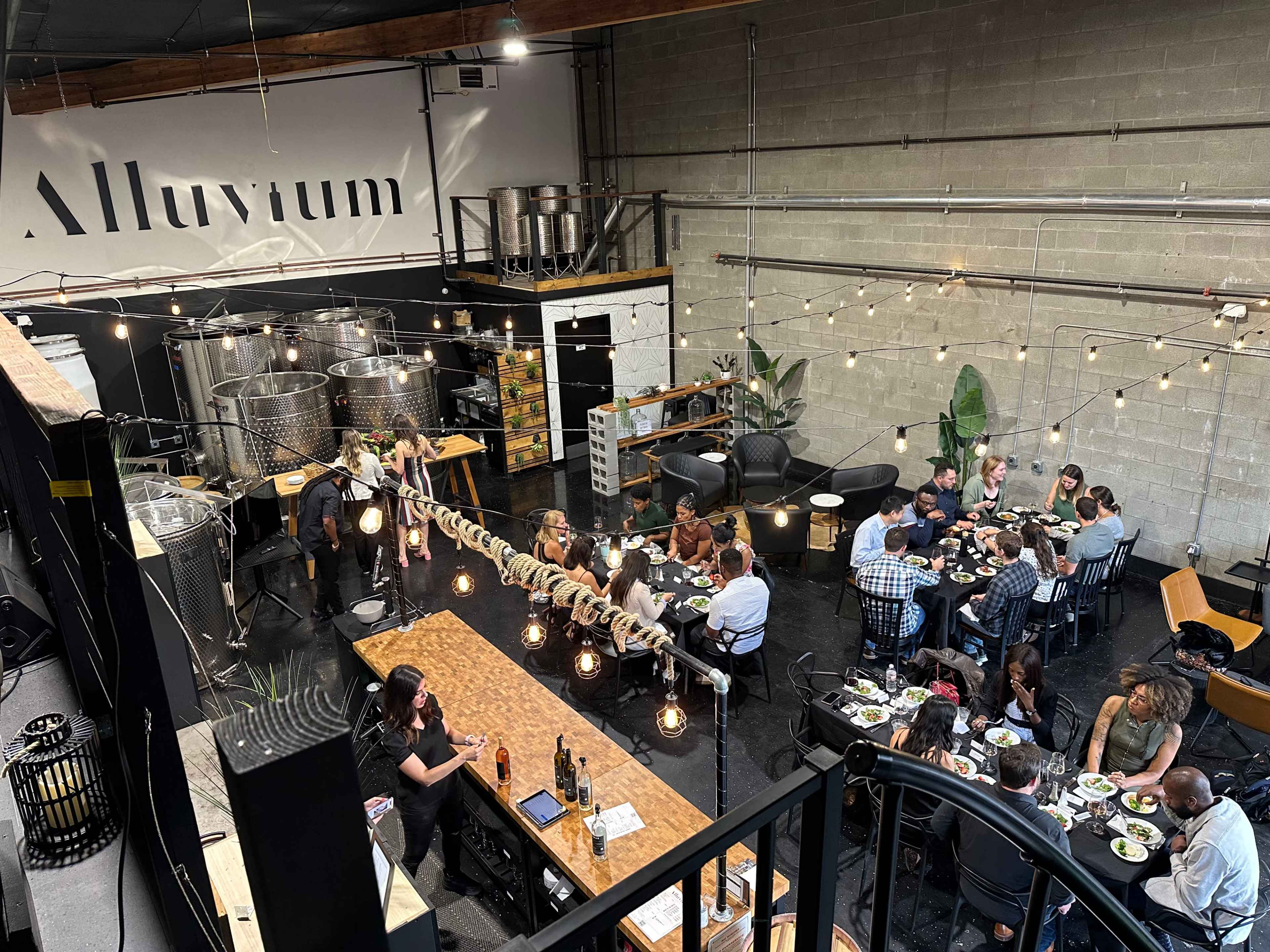 A large group of people gathers around a long wooden table in a spacious venue adorned with metal fermentation tanks and string lights.