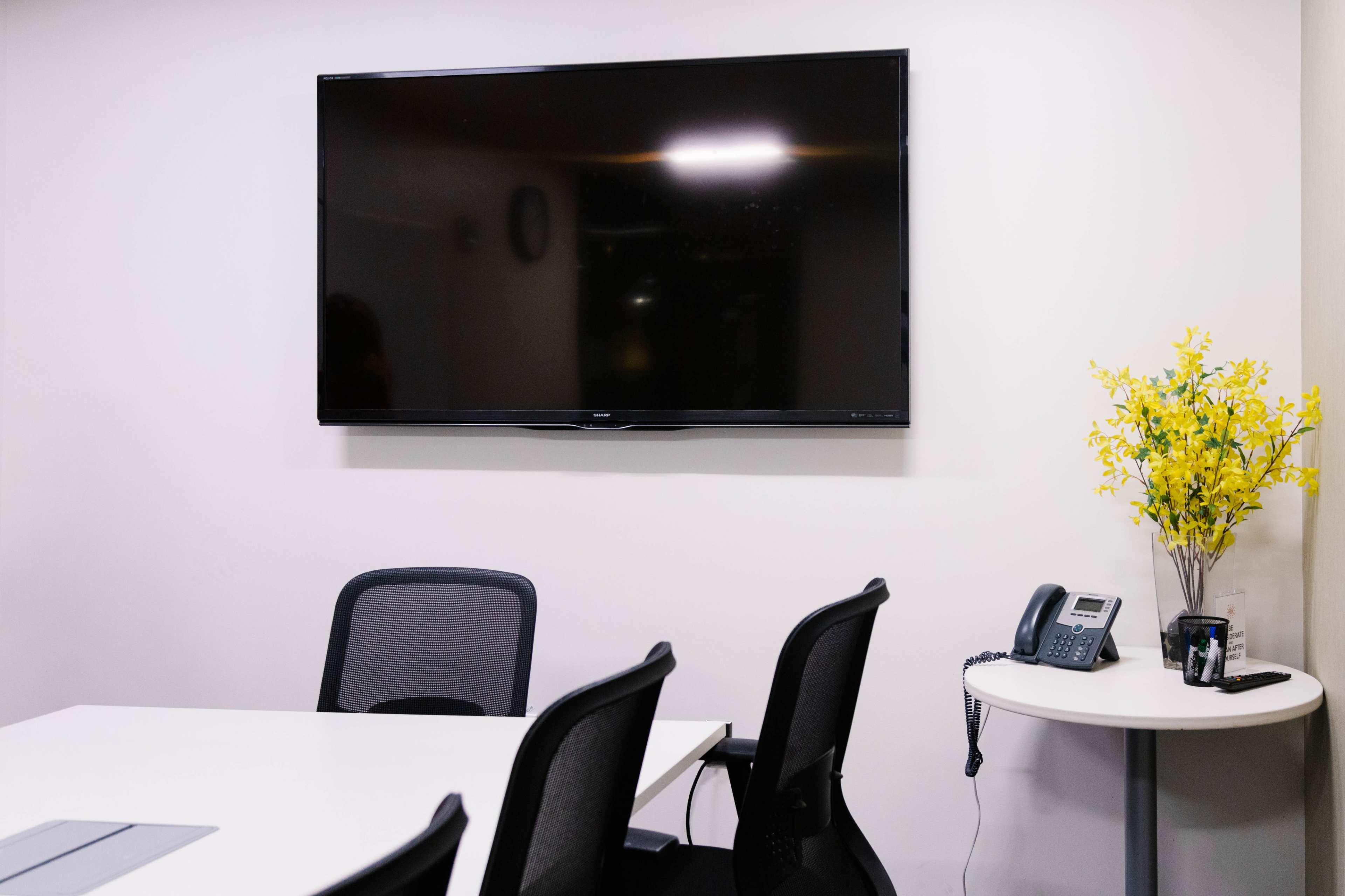 A conference room with a large wall-mounted television, a white table, several black chairs, a phone on a small side table, and a decorative yellow flower arrangement.