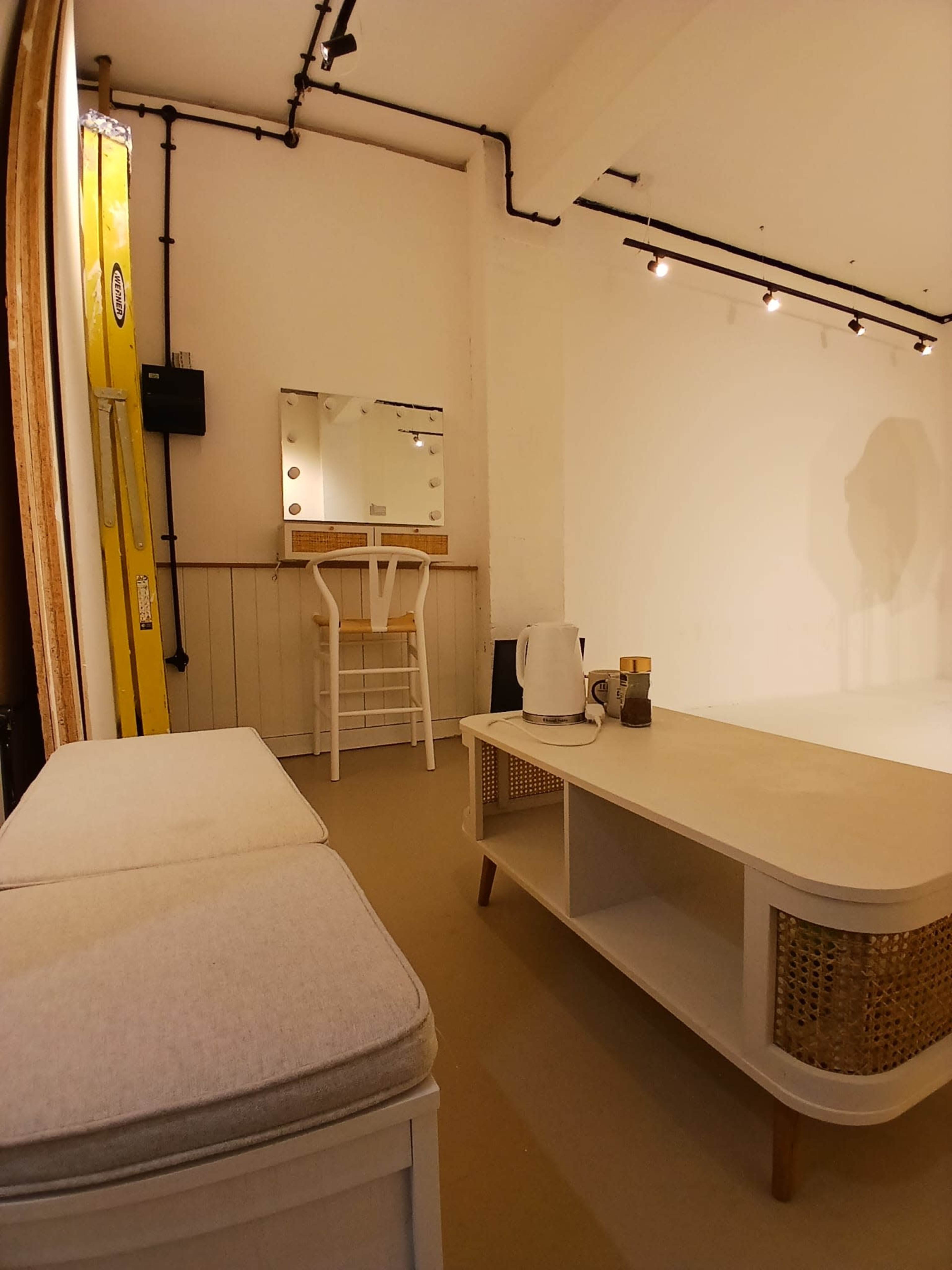 The image shows a minimalist studio space with a small coffee table, a bench, a mirror, and a chair against white walls.