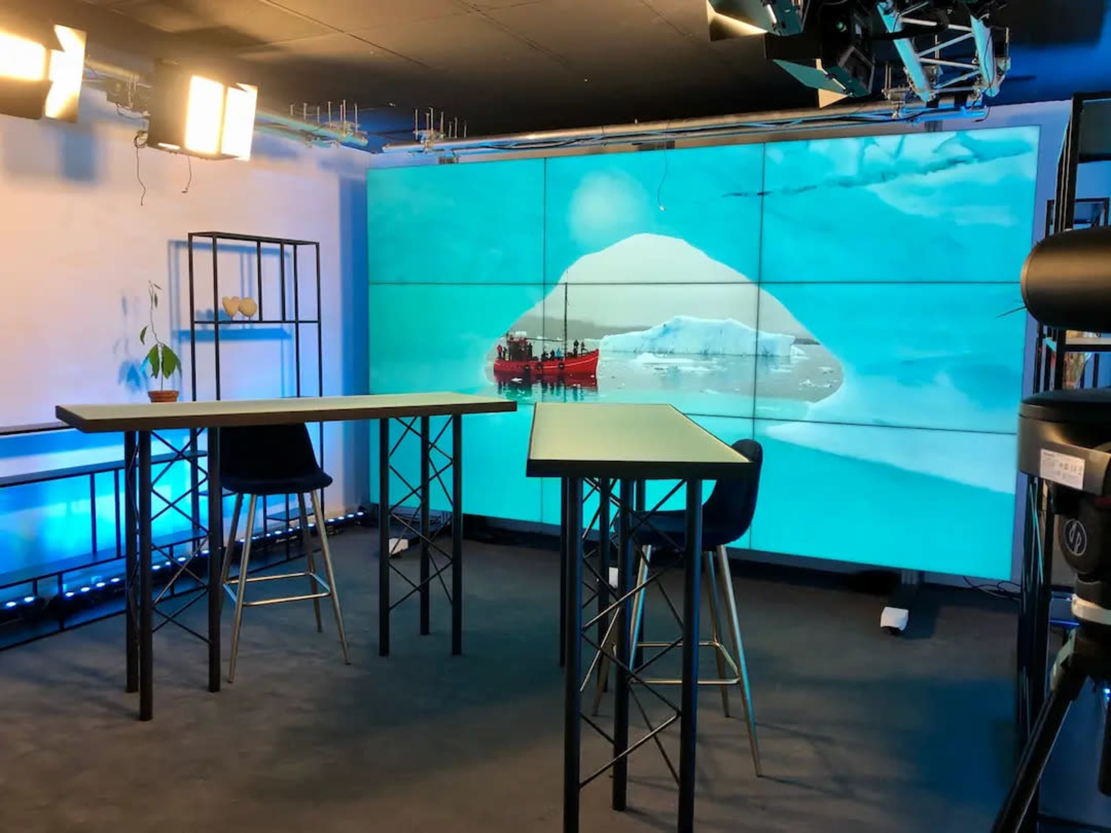 The image shows a modern studio setup featuring two high tables and a large screen displaying an icy landscape with a ship, illuminated by studio lights.