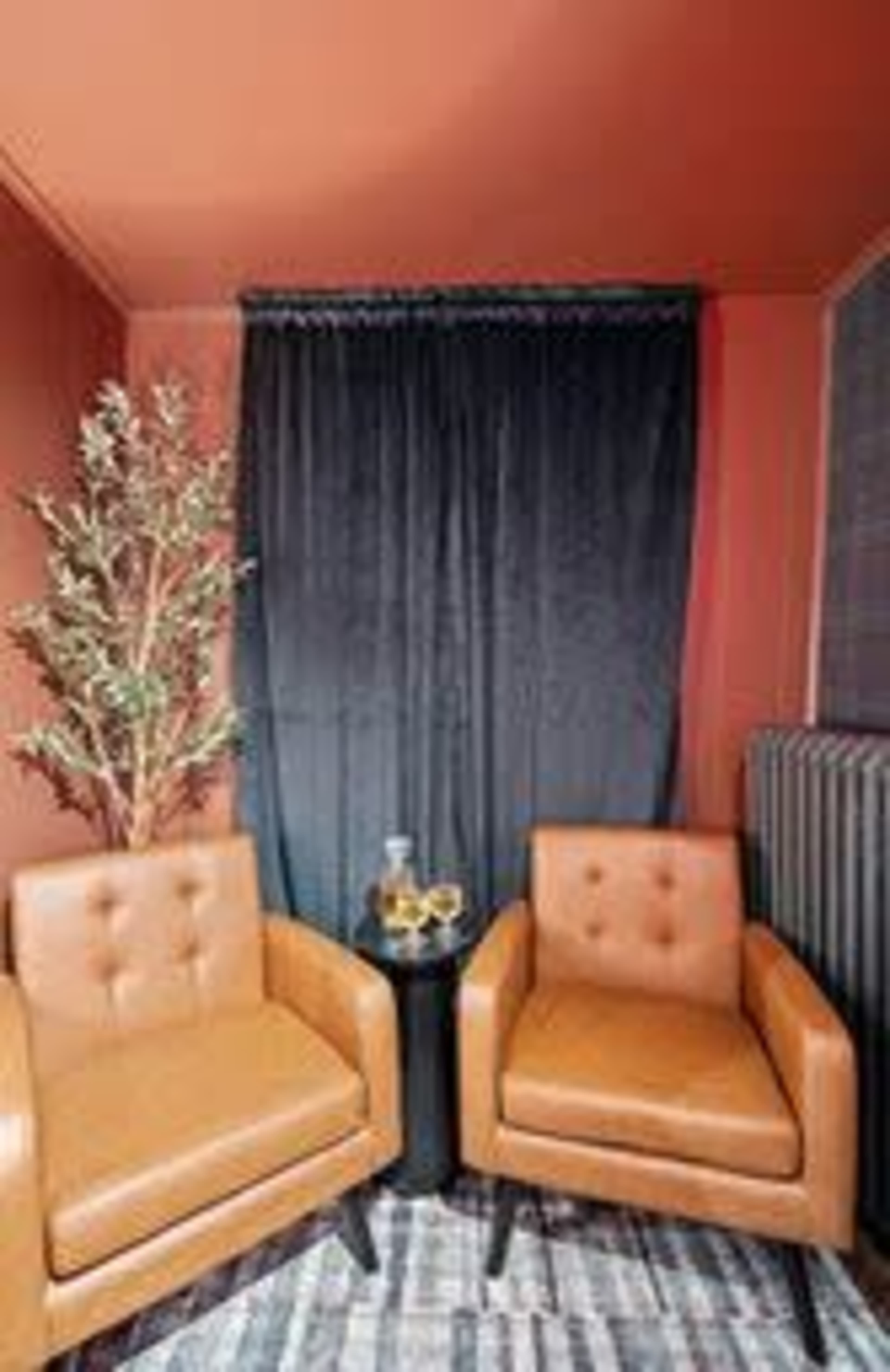 A cozy corner features two brown armchairs, a small black table, a plant, and a dark curtain against a warm-colored wall.