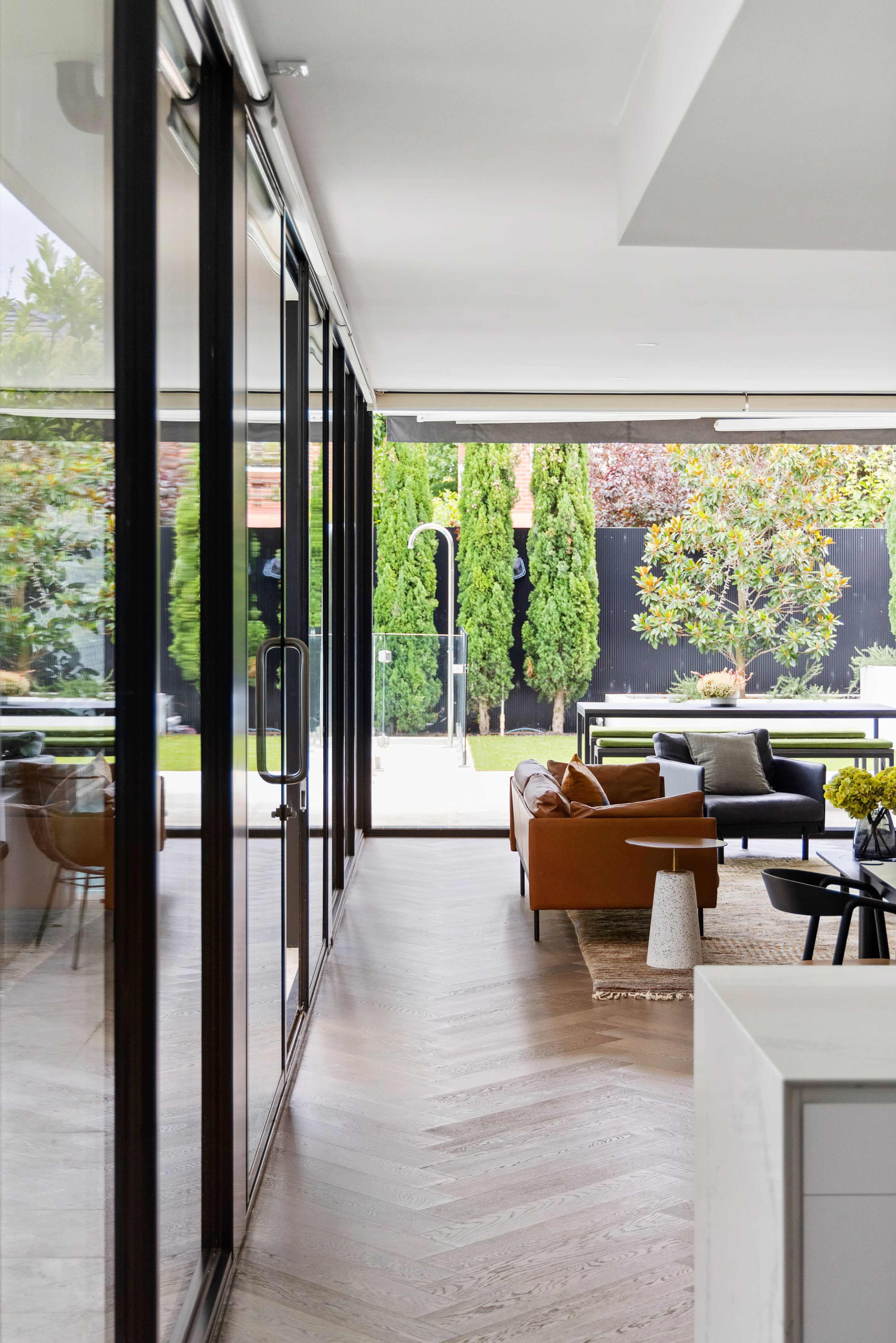 A modern living space features large glass doors that open to a well-landscaped garden with green trees and outdoor seating.