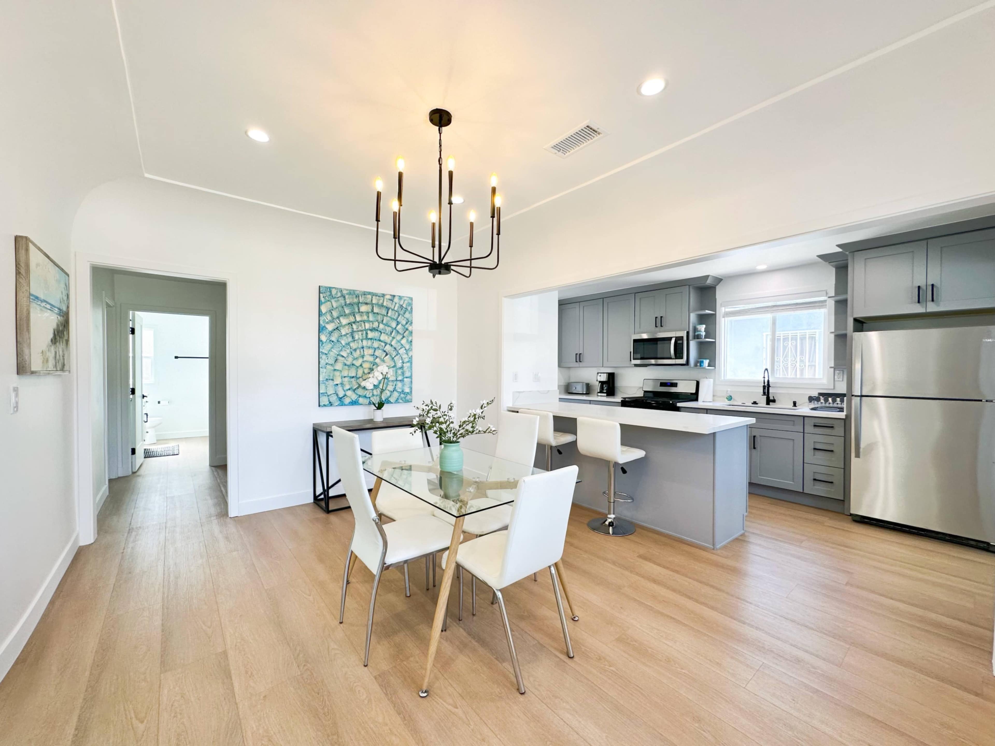 A modern kitchen and dining area features a glass table surrounded by white chairs, with gray cabinetry and stainless steel appliances.