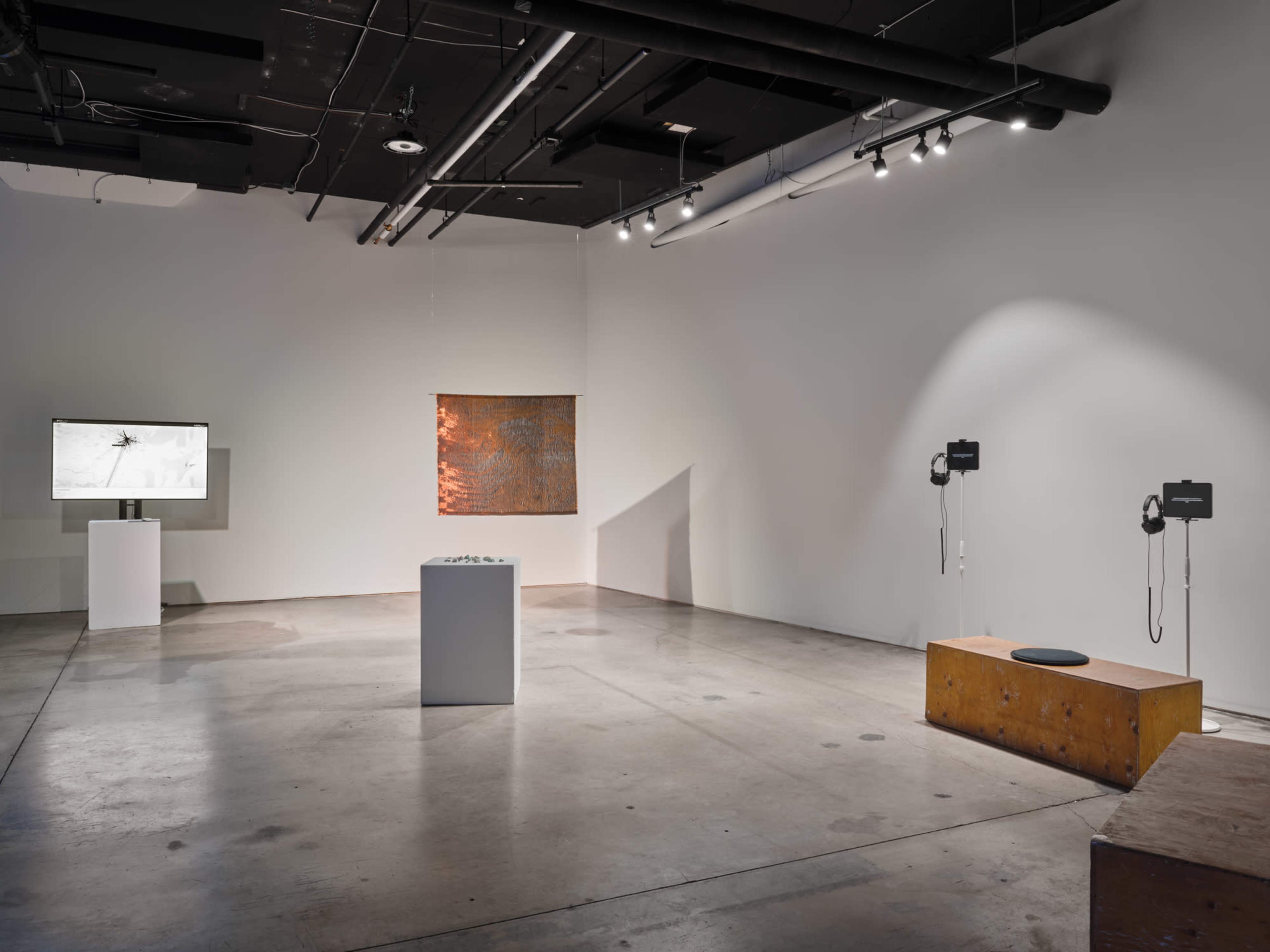 The image shows an art gallery with a minimalist layout featuring an exhibition screen, a central display pedestal, and two installations with headsets on either side.