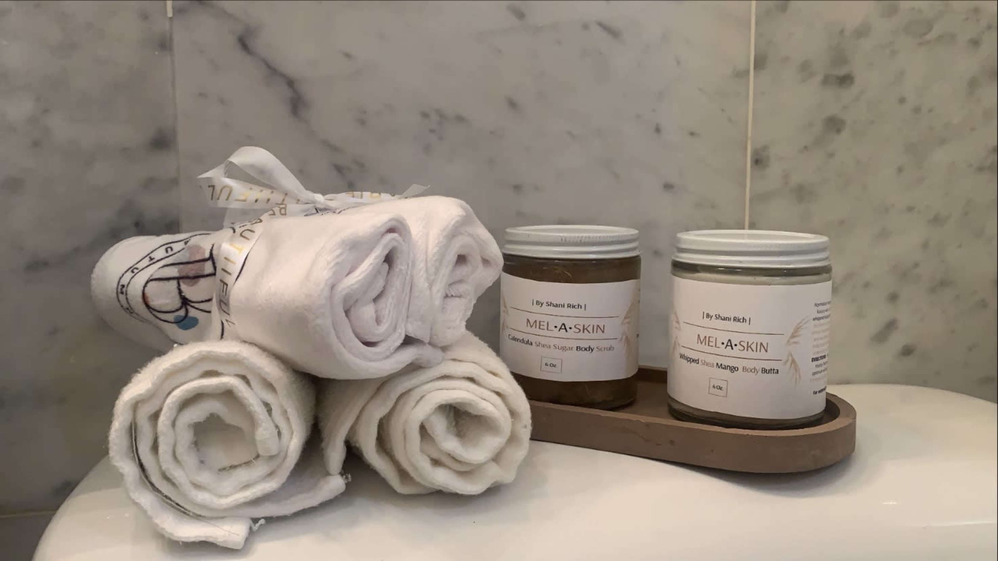 Neatly rolled white towels next to jars of body balm placed on a wooden tray against a marble background.