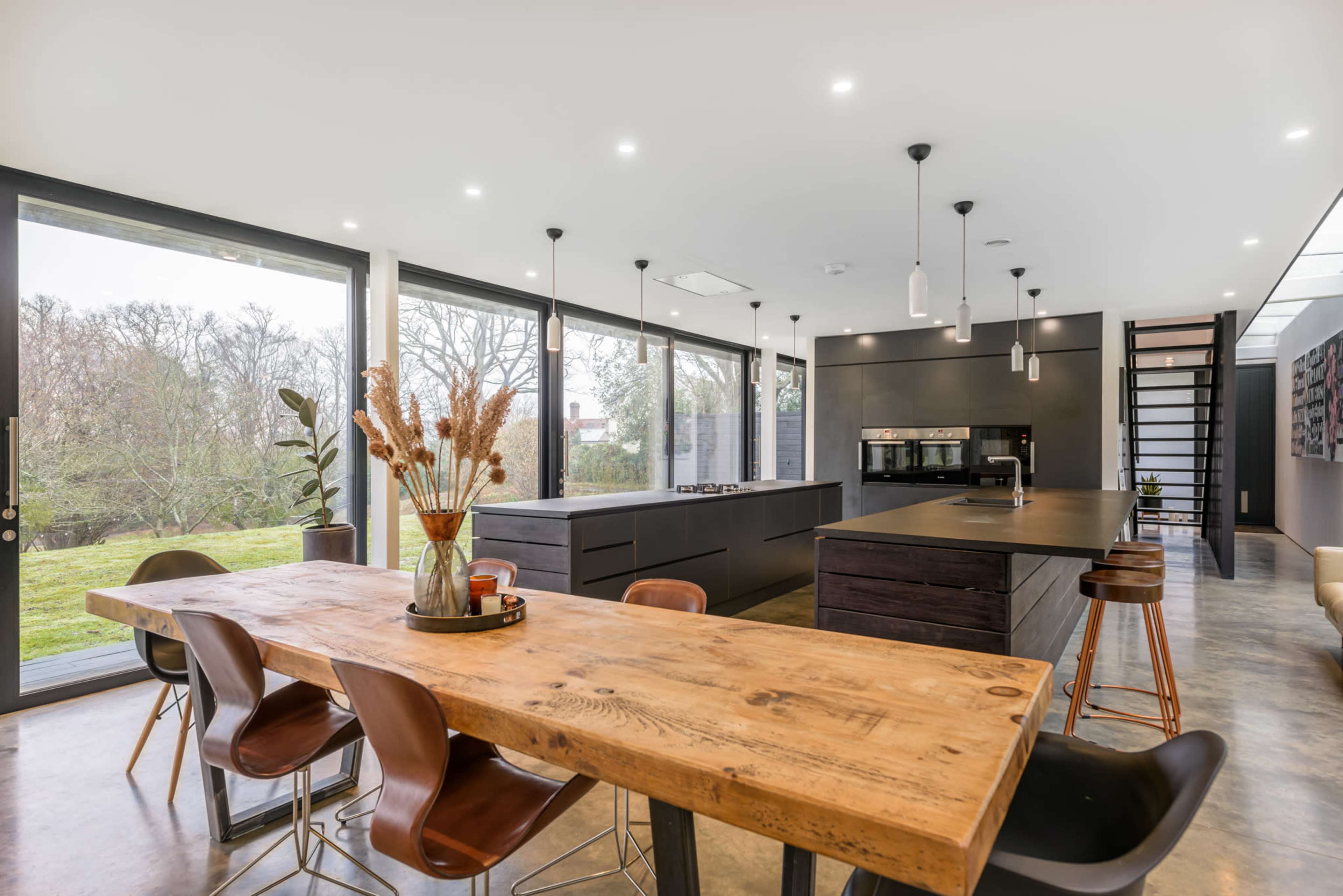 A modern kitchen features a large wooden dining table surrounded by chairs, sleek cabinetry, and large windows overlooking a green landscape.