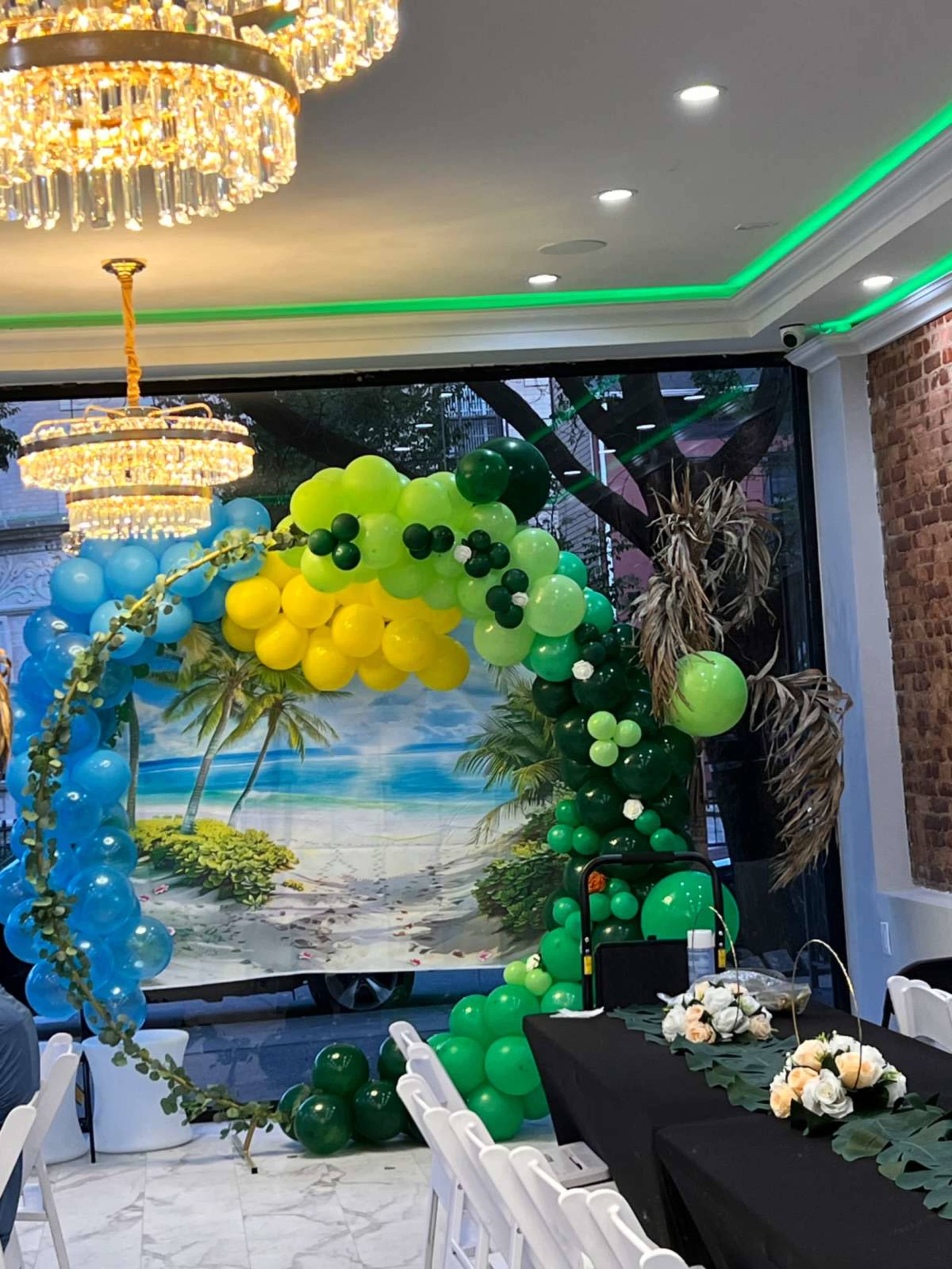 The image shows a decorative arch made of balloons in various shades of blue and green, set against a backdrop of a beach scene, with tables arranged in front.