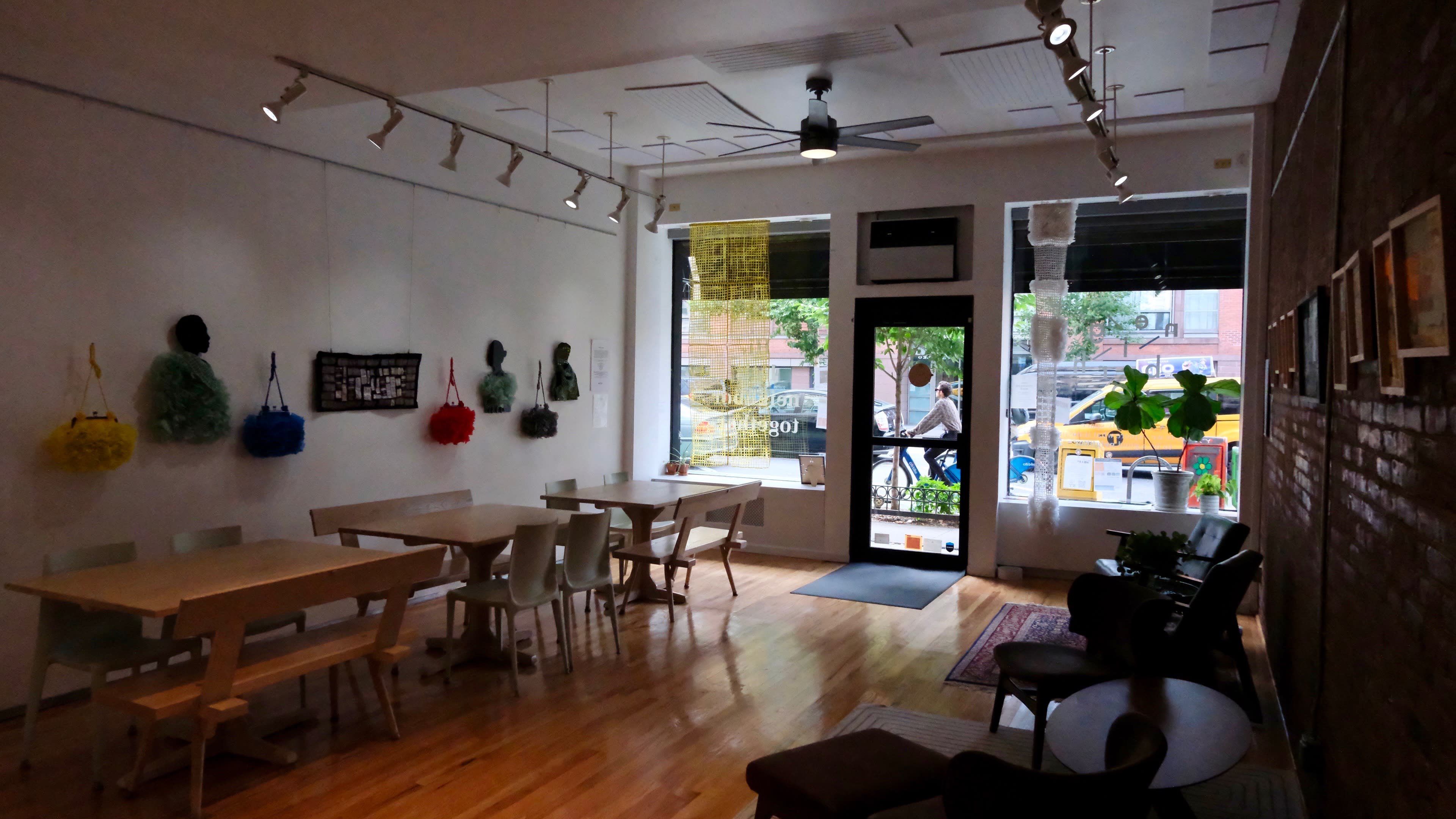 The image shows a modern cafe interior with wooden tables, chairs, and colorful artworks displayed on the walls, along with large windows facing a street.
