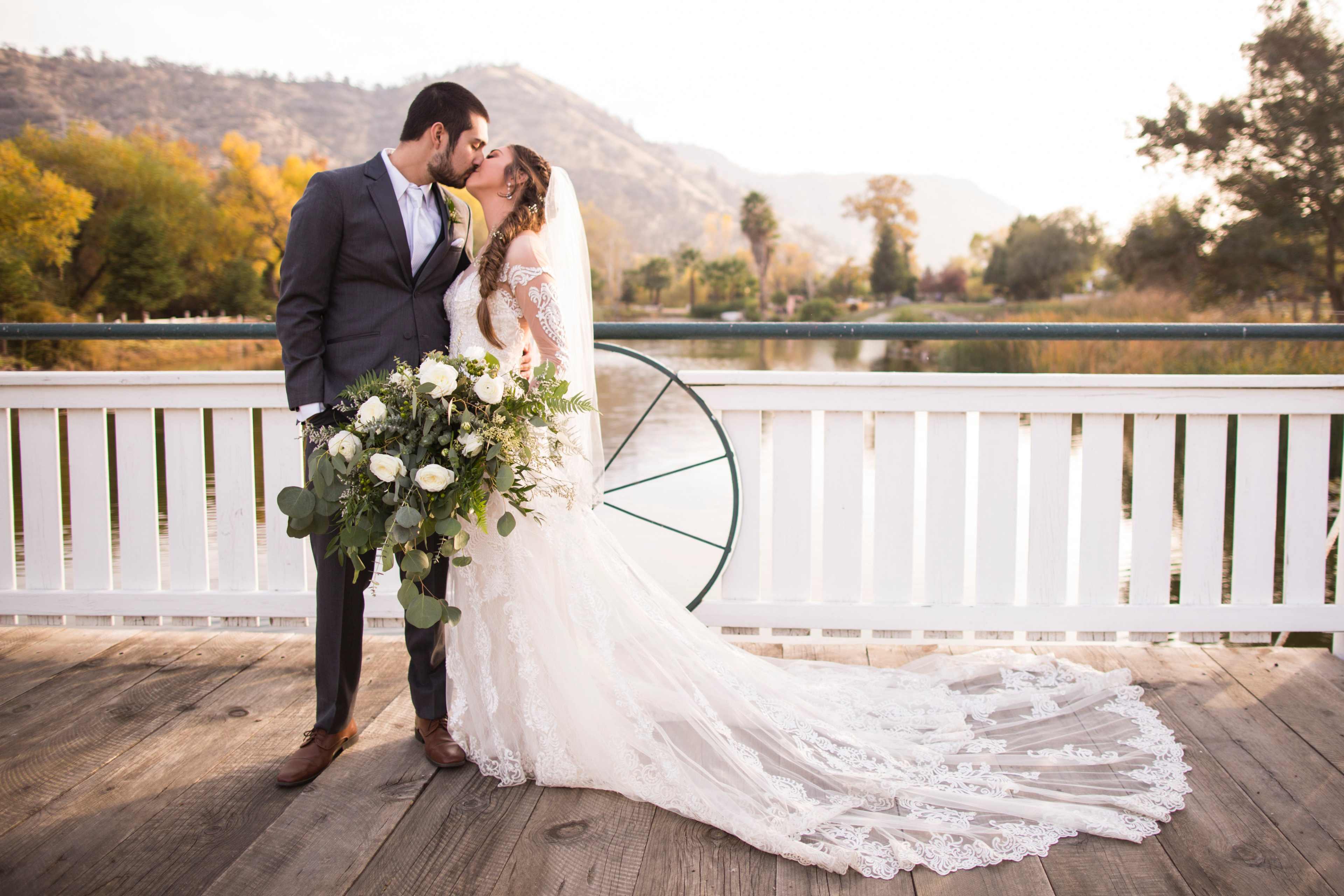 A bride and groom share a kiss on a wooden dock by a serene body of water, surrounded by autumn foliage.