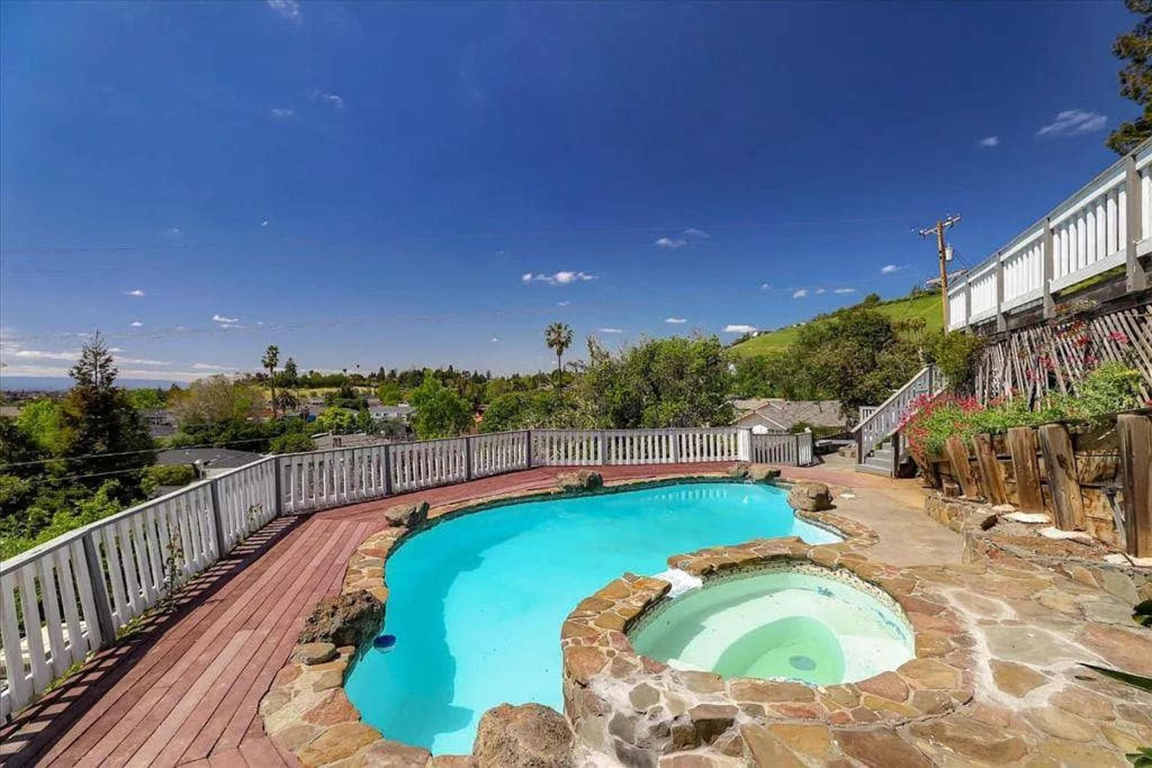 The image shows a backyard with a curved swimming pool and a hot tub, surrounded by a stone patio and lush greenery under a clear blue sky.