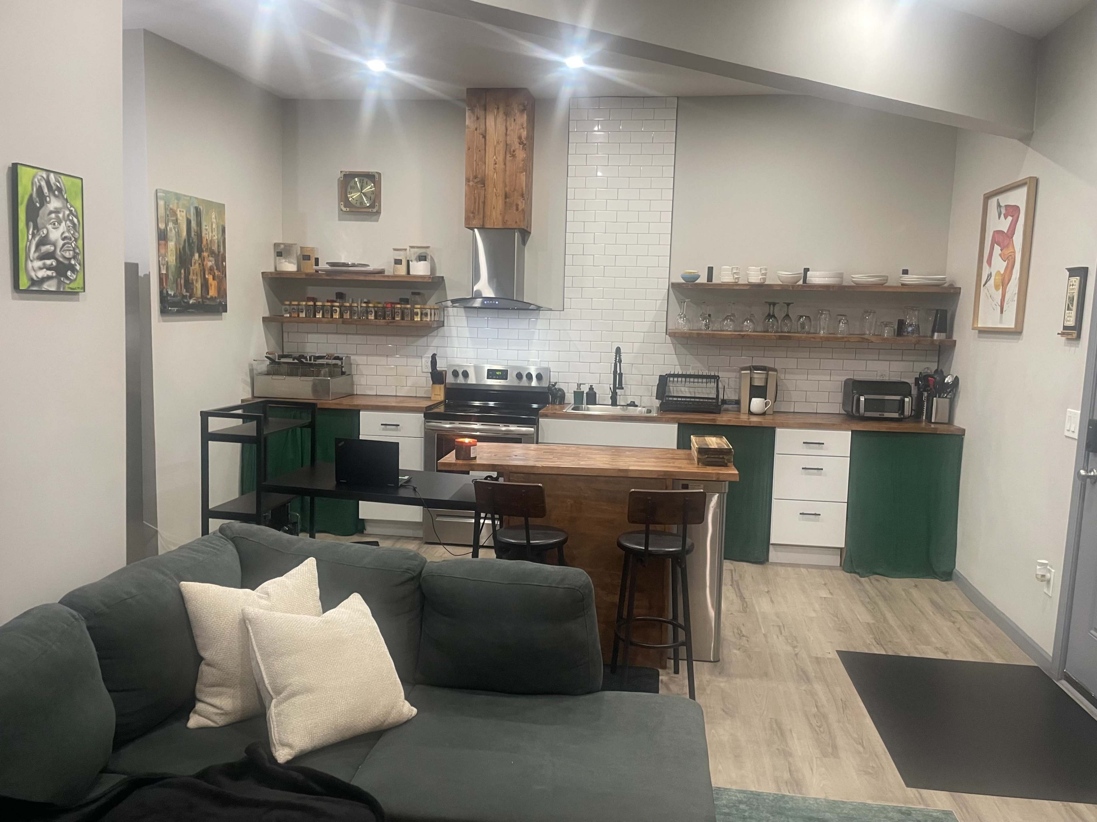 The image shows a modern kitchen with a wooden countertop, stainless steel appliances, and shelves holding kitchenware, adjacent to a cozy living area featuring a green sofa and decorative cushions.