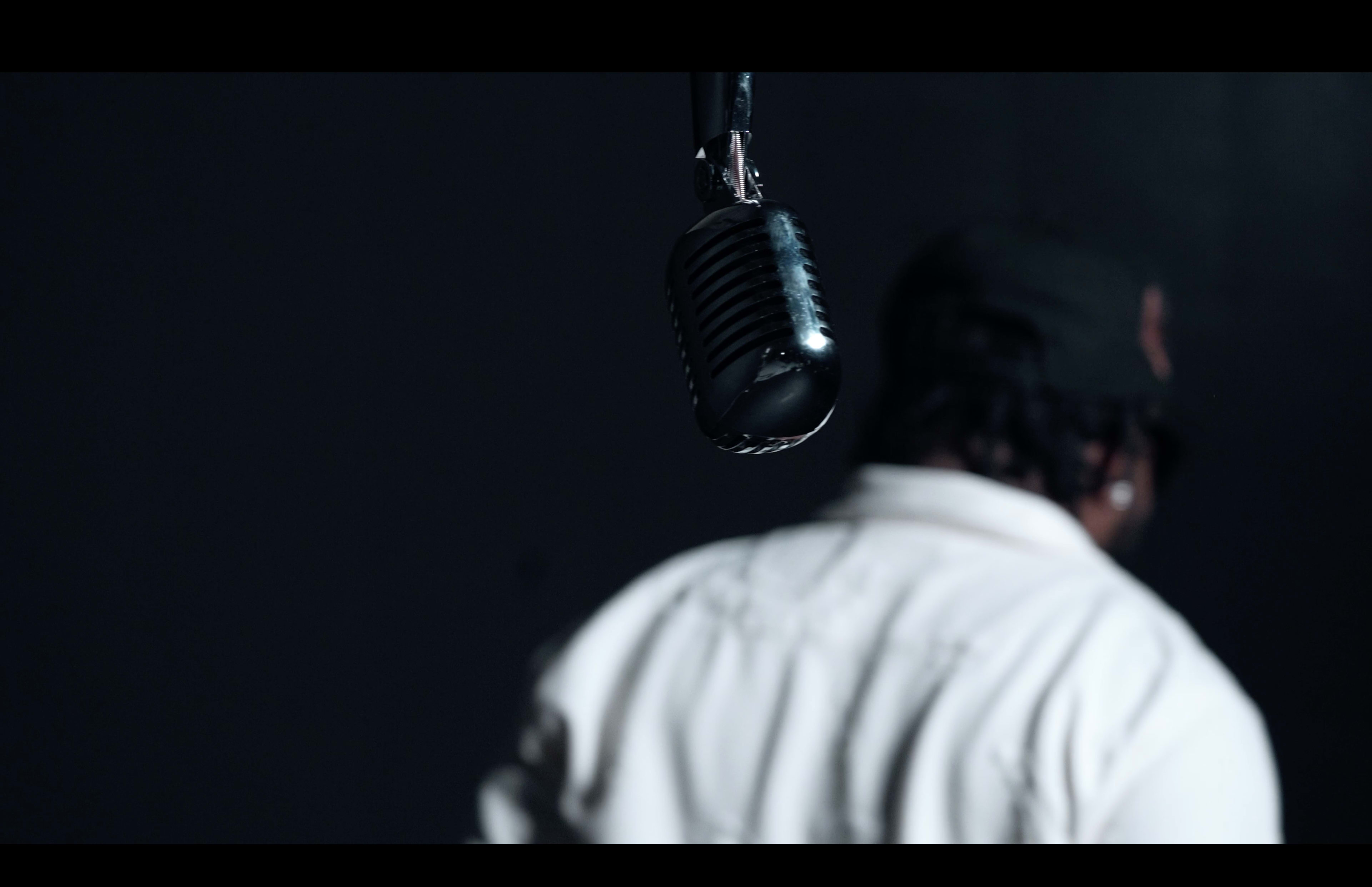 A vintage microphone hangs from above in focus while a person with their back turned, wearing a cap, stands in the background.