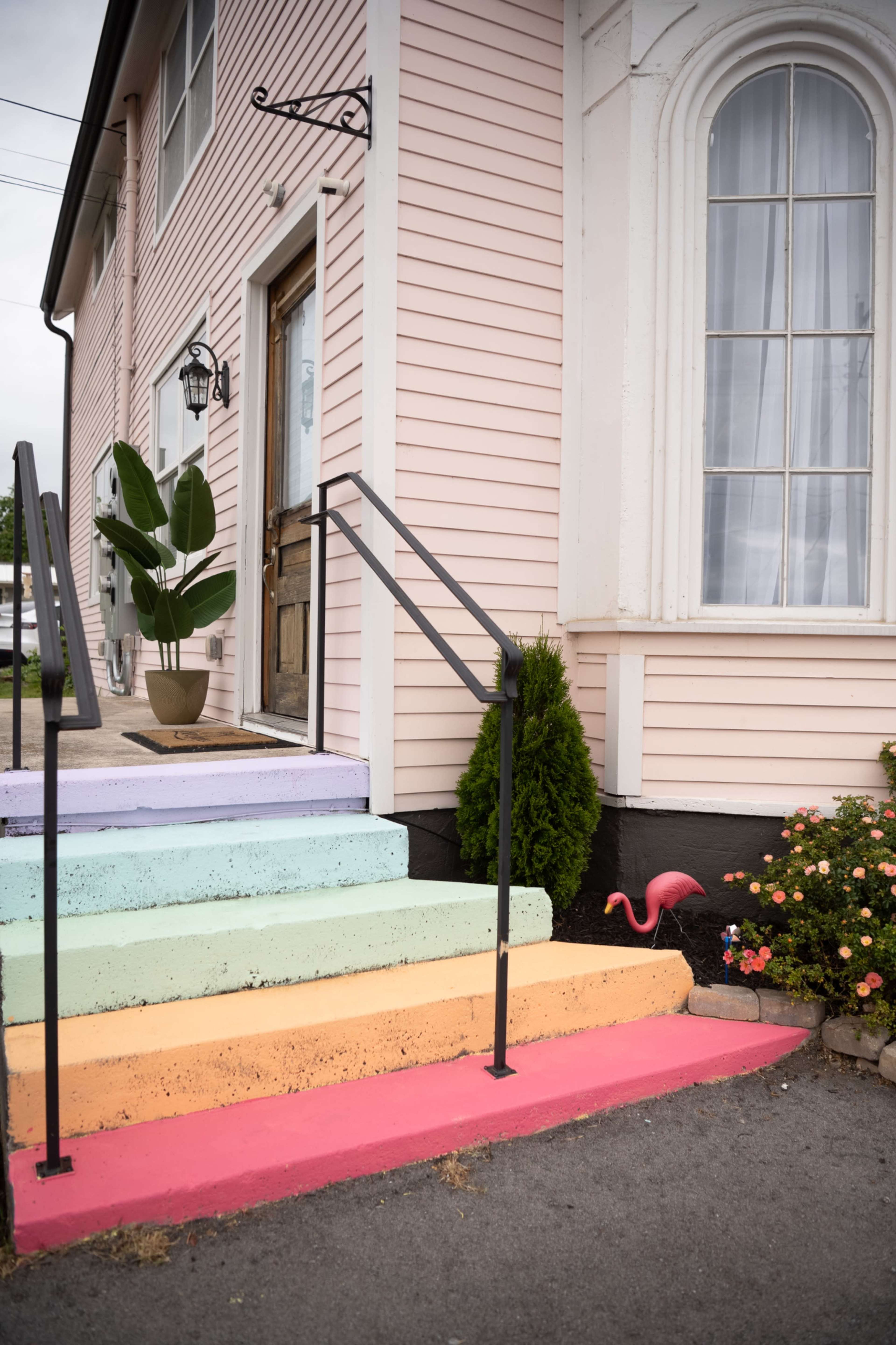 The image shows a pastel-colored staircase leading to a building entrance, with a potted plant nearby and a garden with flowers and a pink flamingo decoration.